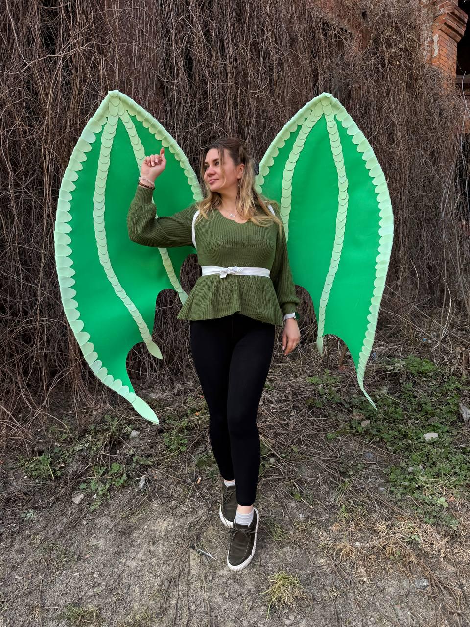 May include: A person wearing large, bright green dragon wings with a scalloped edge. The wings are attached to a white harness. The person is wearing a green sweater, black leggings, and dark green sneakers.