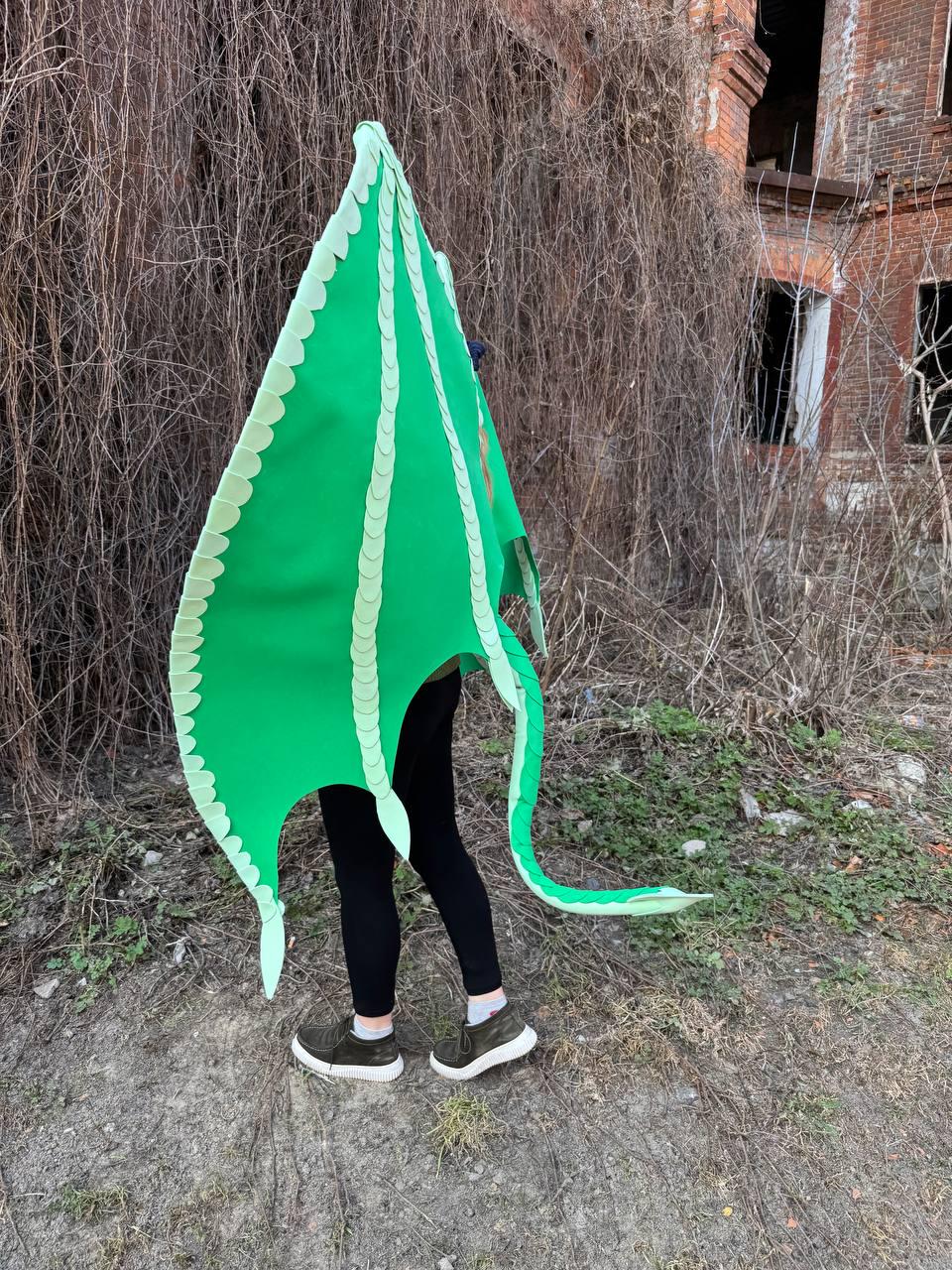 May include: A bright green dragon wing costume with a scalloped edge and a long tail. The costume is worn by a person wearing black leggings and dark green sneakers. The background features overgrown foliage and a brick building.