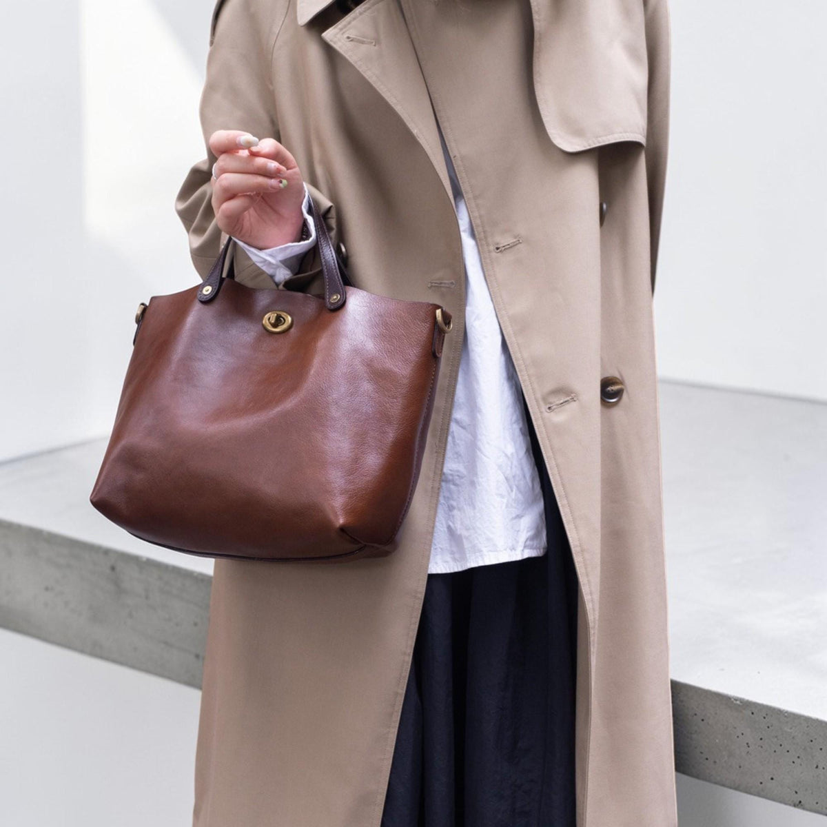 May include: A brown leather tote bag with a top handle and a gold-colored clasp. The bag is held by a person wearing a tan trench coat and a white shirt. The bag has a classic, minimalist design.