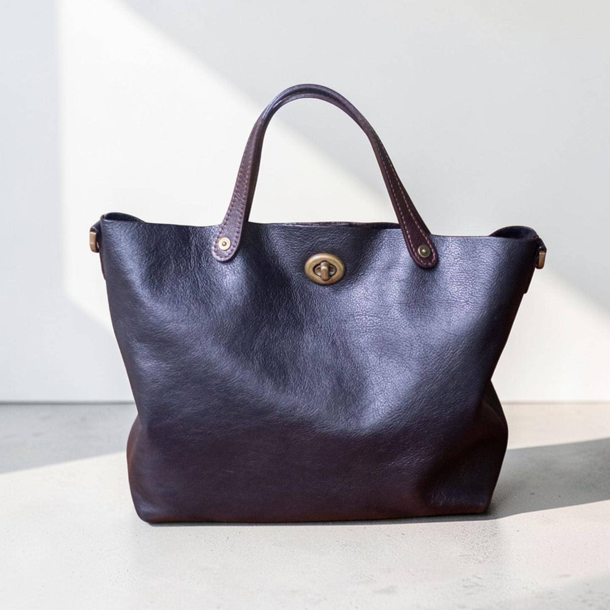 May include: A dark brown leather tote bag with a structured design. The bag features a top handle with brown accents, a gold-colored clasp, and a spacious interior. The bag is designed for everyday use.