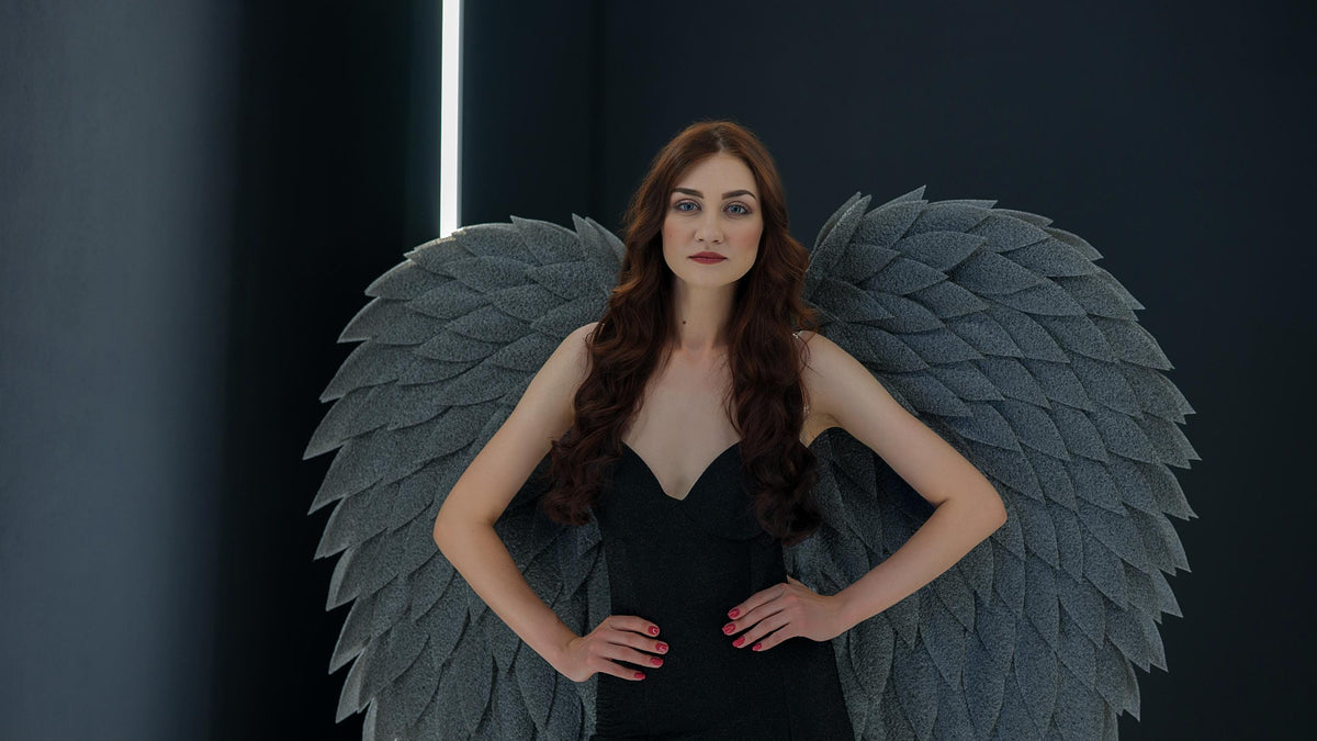 May include: A woman wearing a black dress and large gray feathered wings. The wings are a prominent feature, with a layered design. The woman has long, wavy auburn hair and is looking directly at the camera.