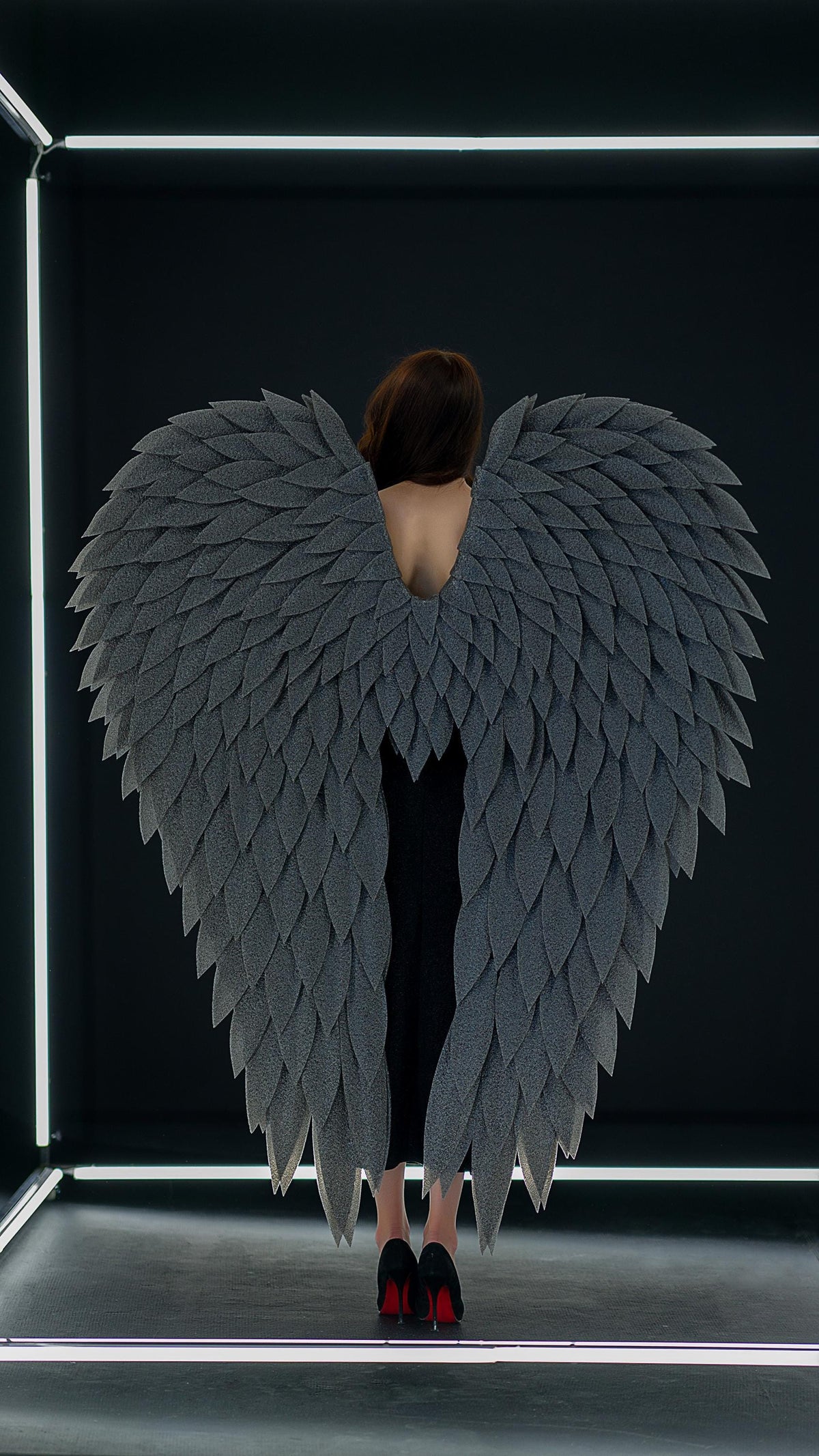 May include: A person wearing large, gray, feathered angel wings. The wings are the focal point, with a layered design. The person is wearing a black dress and black heels with red soles. The background is black with white neon lights.