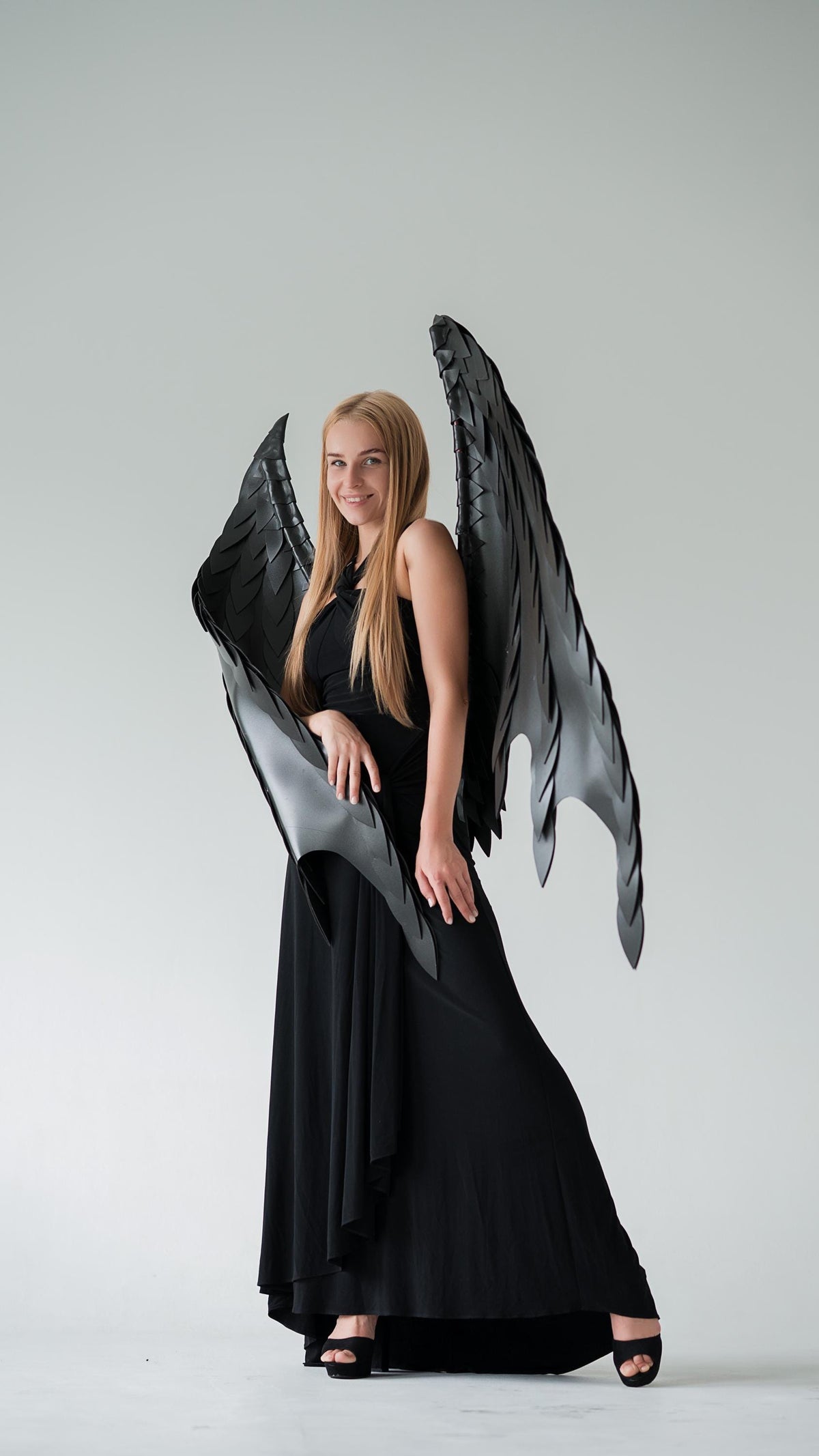 May include: A woman wearing a black dress and large, black, feathered angel wings. The wings are detailed with layered segments, and the dress is a flowing, floor-length gown. The background is a plain, light gray.