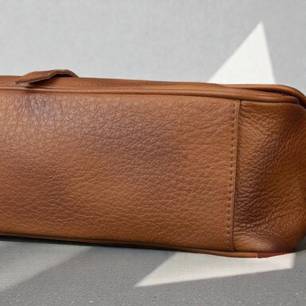 May include: A close-up of a brown leather handbag. The bag has a rectangular shape with a flap closure and a textured surface. The stitching is visible along the edges, and the leather has a natural, worn appearance.