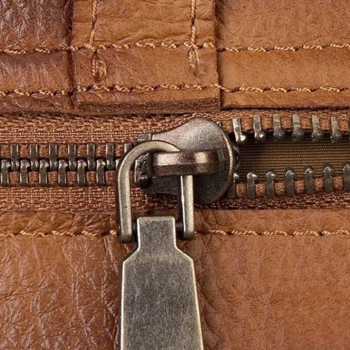 May include: Close-up of a brown leather zipper. The zipper pull is a silver-toned metal, and the leather has visible stitching. The zipper teeth are also silver-toned, and the leather has a textured appearance.