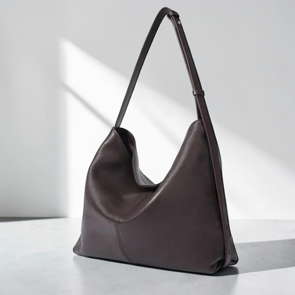 May include: A dark brown leather shoulder bag with a long strap. The bag has a curved shape and a minimalist design. The bag is made of a smooth leather material.