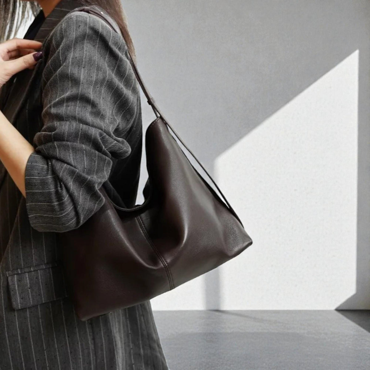 May include: A dark brown leather shoulder bag with a long strap. The bag has a simple, minimalist design and is worn over the shoulder. The bag is made of a smooth, supple leather.