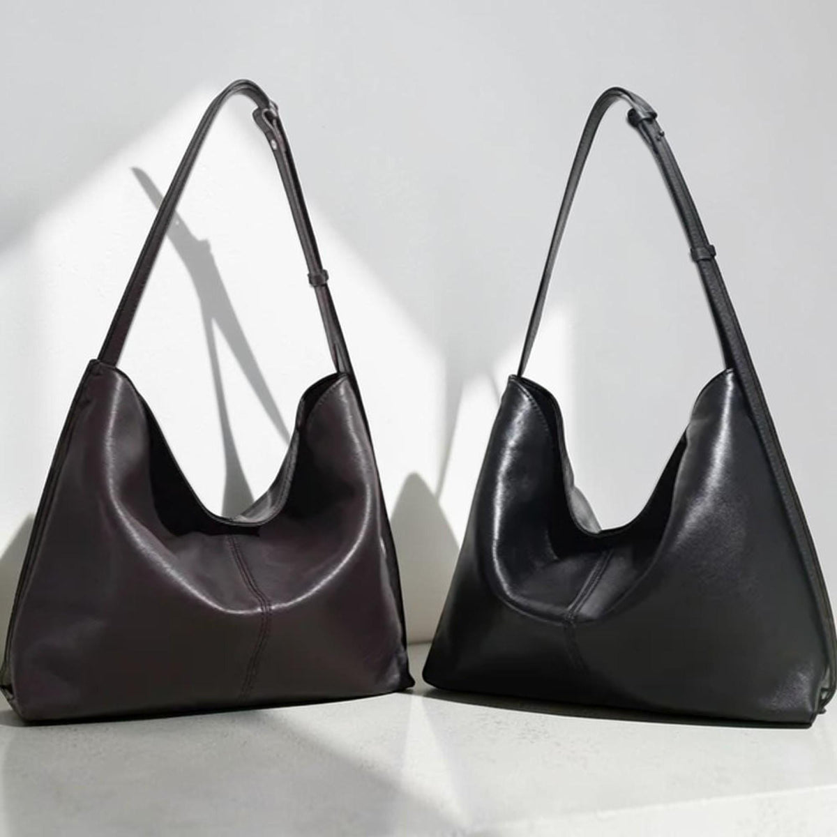 May include: Two leather shoulder bags. One is a deep burgundy color, and the other is black. Both bags have a crescent shape with a long, thin strap. The bags are displayed against a white background.