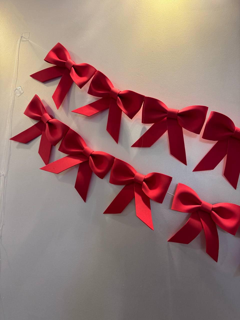 May include: A string of vibrant red bows, each meticulously tied, are suspended against a neutral-toned wall. The bows are evenly spaced, creating a festive and decorative garland. The bows are made of a smooth, satin-like material.