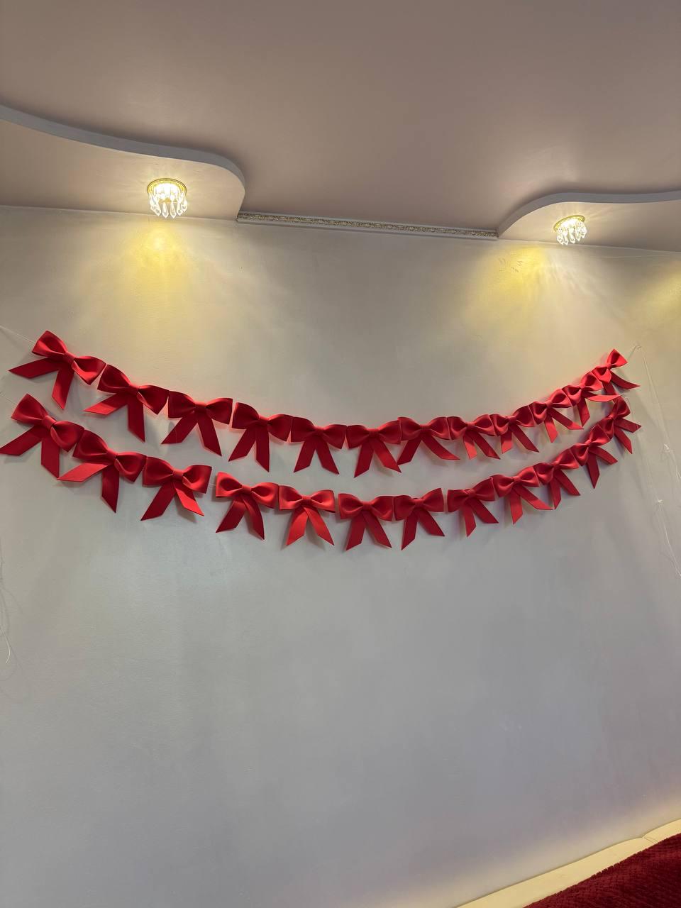May include: Two rows of bright red bows are strung across a white wall. The bows are evenly spaced and appear to be made of a satin-like material. The wall is lit by two small, ornate ceiling lights.