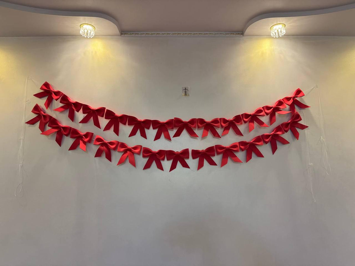 May include: Two rows of red satin bows are strung across a white wall. The bows are evenly spaced and appear to be part of a decorative garland. Two small lights are mounted on the ceiling above the bows.