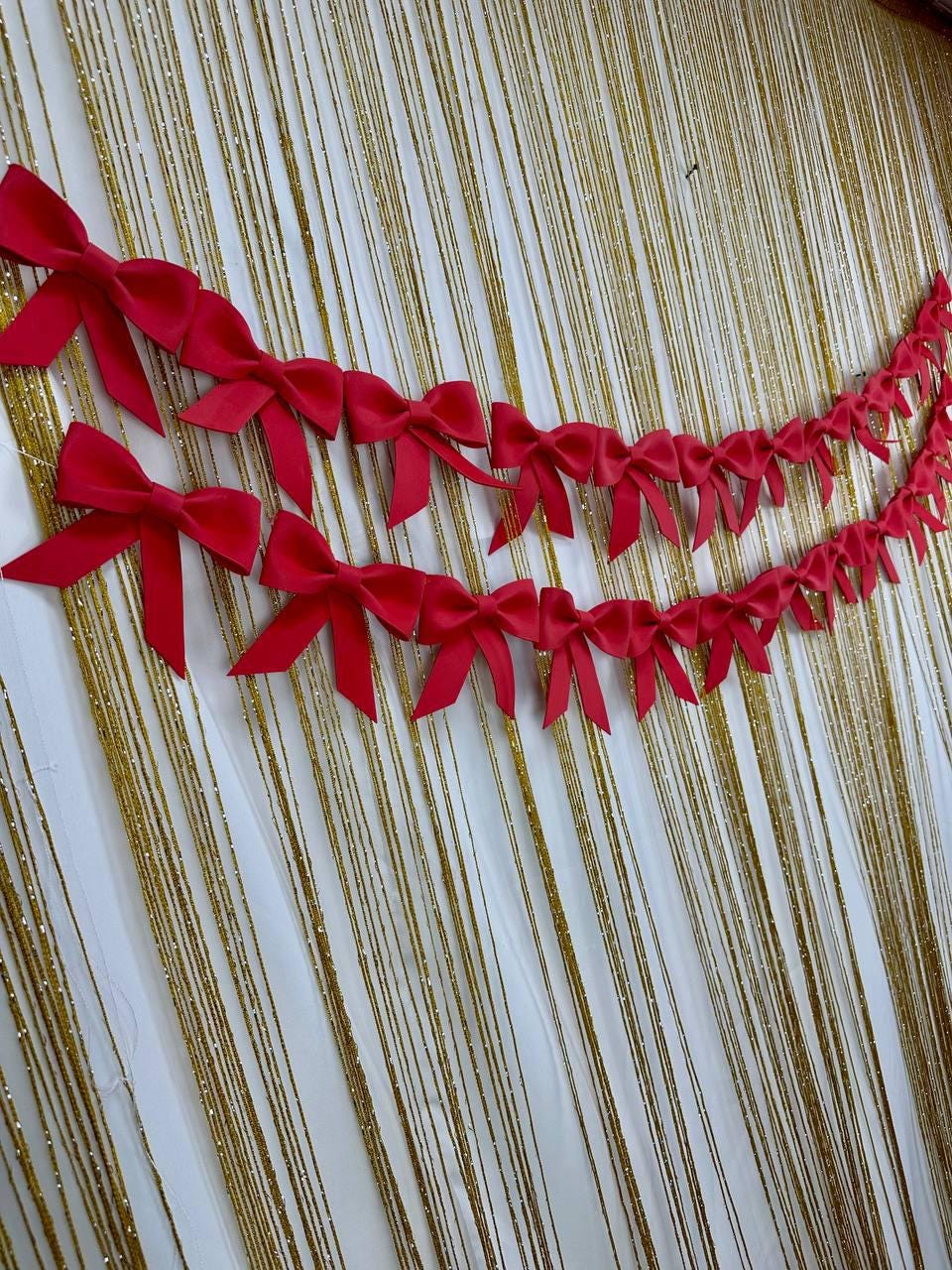 May include: Two strands of bright red satin bows are arranged in a decorative garland. The bows are set against a backdrop of gold tinsel strands, creating a festive and eye-catching display.
