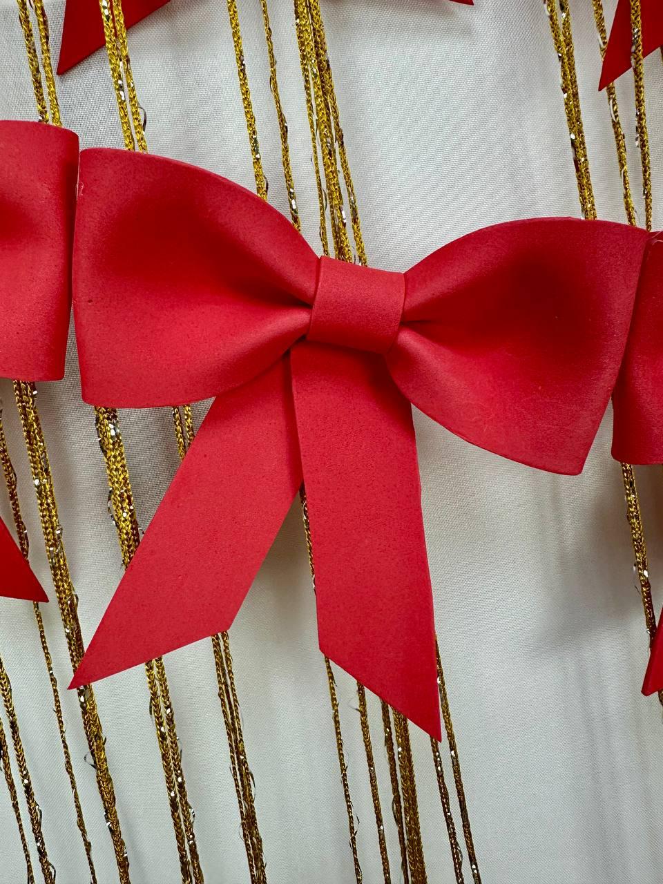 May include: Close-up of a red bow with long tails, part of a decorative garland. The bow is made of a matte material and is set against a backdrop of gold strands and a white surface. Festive decor.