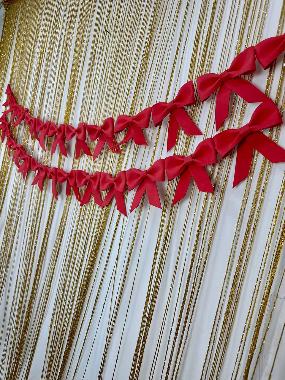 May include: Two strands of bright red satin bows are suspended against a backdrop of gold and white shimmering strands. The bows are evenly spaced and create a festive, decorative element. The background has a vertical, textured pattern.