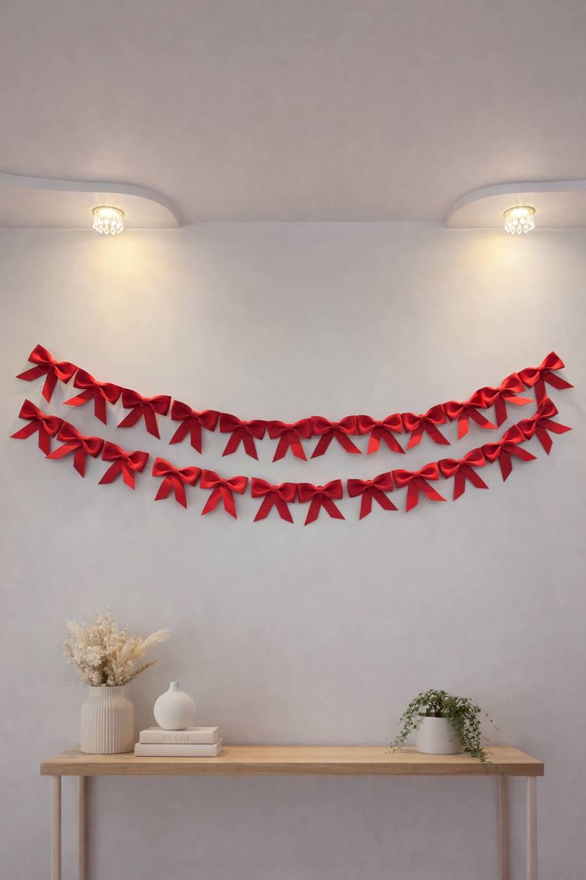 May include: Two rows of bright red satin bows are arranged in a decorative garland on a white wall. The bows are evenly spaced and create a festive, eye-catching display. The wall is lit by two small ceiling lights.
