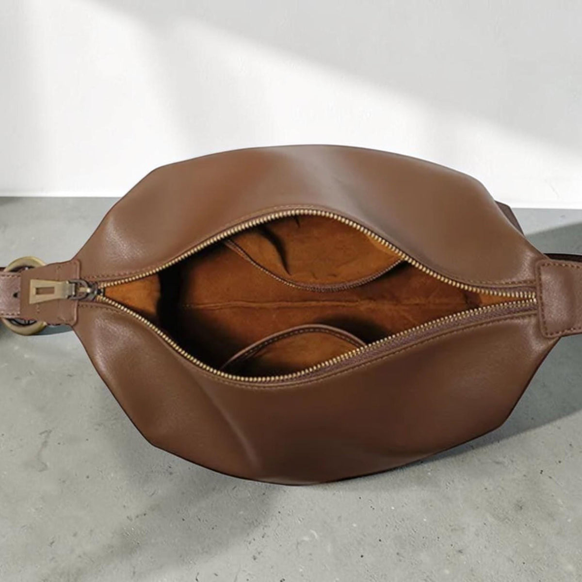 May include: A brown leather hobo bag with a wide, open top and a gold-toned metal zipper closure. The interior is lined with a soft, brown suede-like material and features a central divider, creating two distinct compartments within the bag.