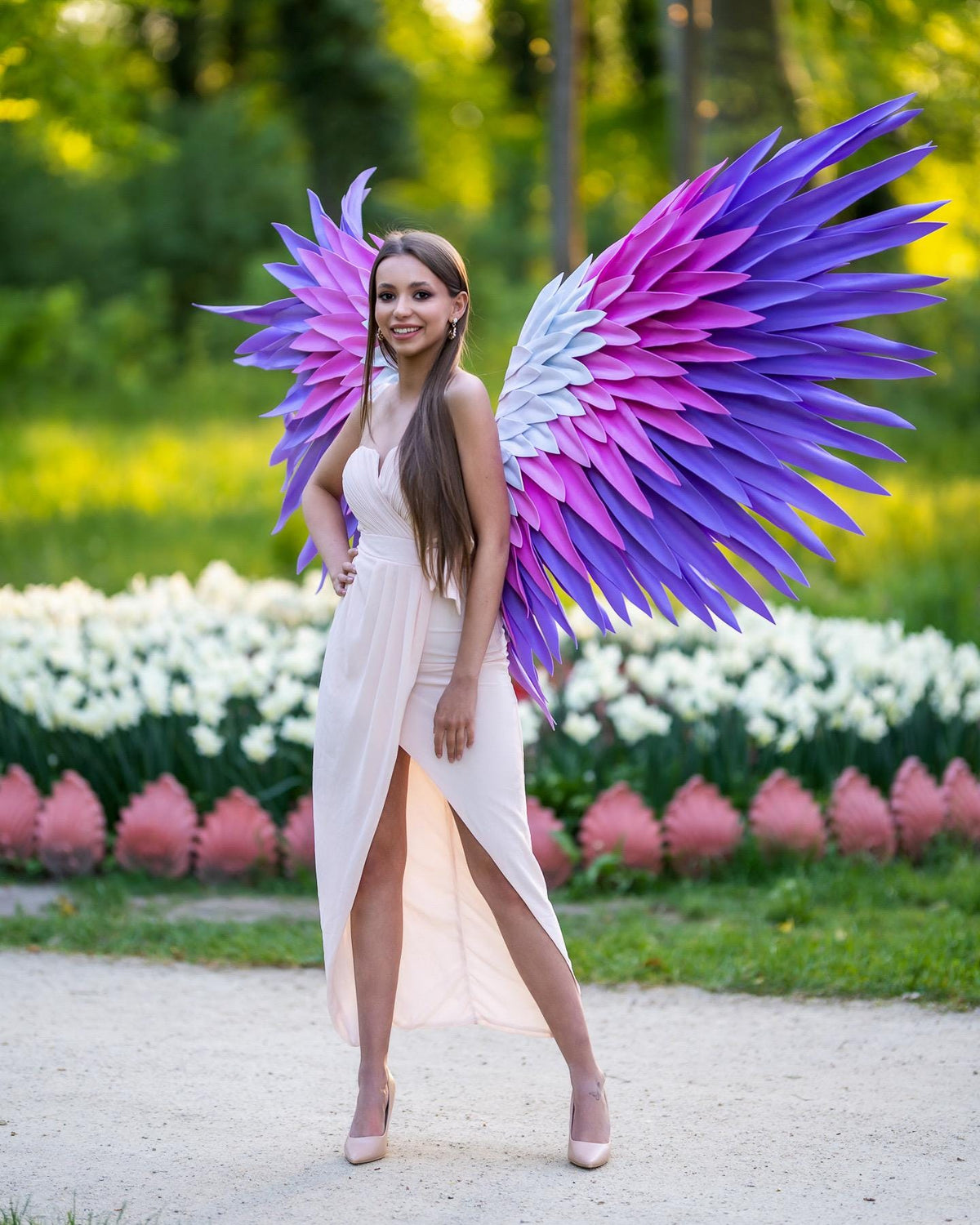 May include: A pair of large, decorative wings featuring a gradient of purple, magenta, and white feathers. The wings are attached to a light beige, sleeveless, high-low dress with a sweetheart neckline and a draped, wrap-style skirt design.