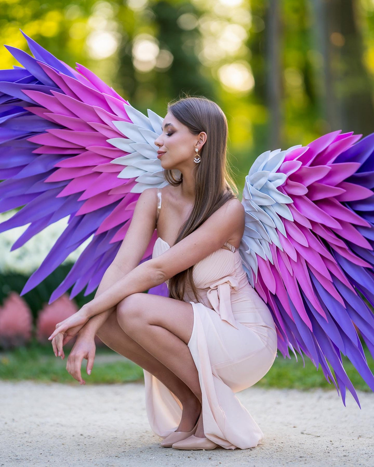 May include: Large decorative wings featuring layered, pointed feathers in a gradient of purple, magenta, and white. The wings are constructed with a structured frame and individual feather-like segments that extend outward in a wide, expansive arc.