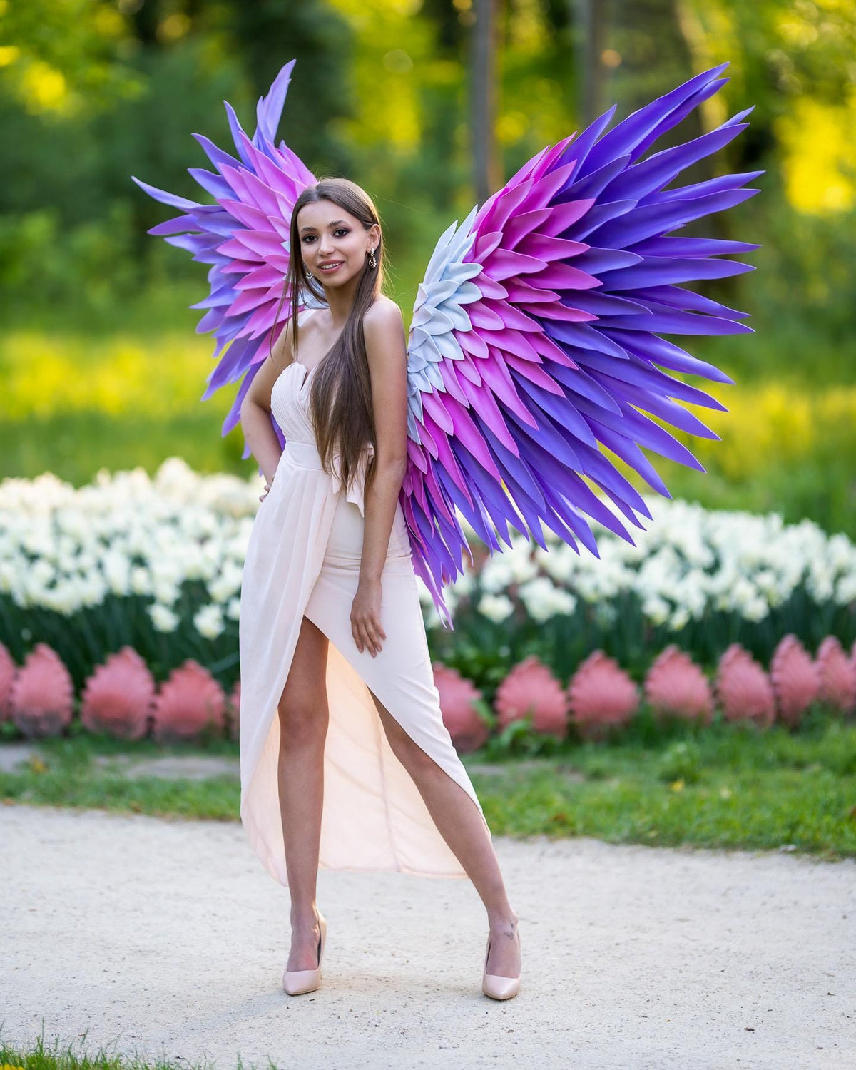 May include: Large decorative angel wings featuring a layered feather design in a gradient of purple, magenta, and light blue. The wings are worn as a costume accessory, showcasing pointed, rigid feather-like structures that extend outward in a wide span.