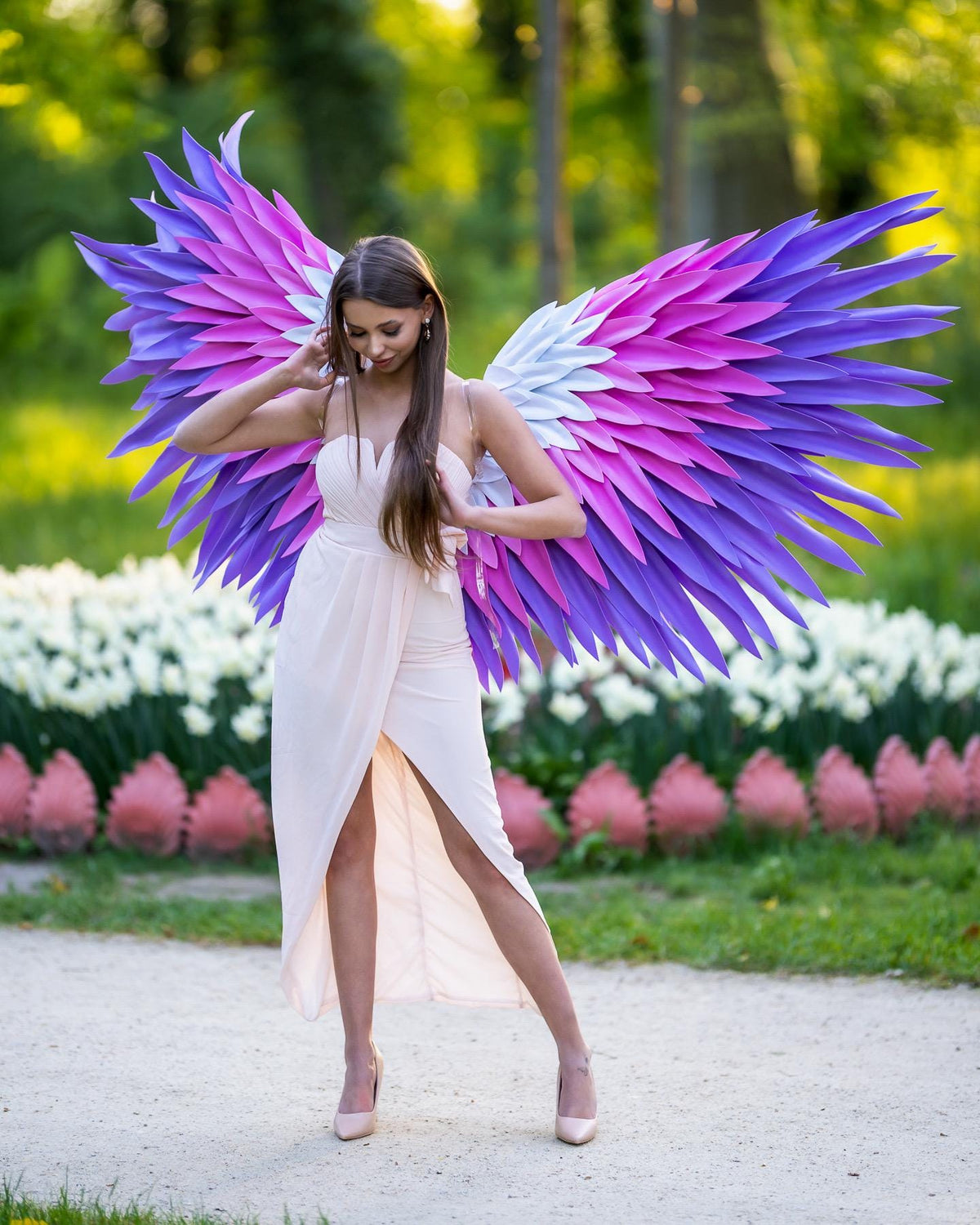 May include: Large decorative wings featuring layered feathers in shades of purple, magenta, and white. The wings are attached to a light beige sleeveless dress with a wrap-style skirt and a high front slit, creating a layered and textured appearance.