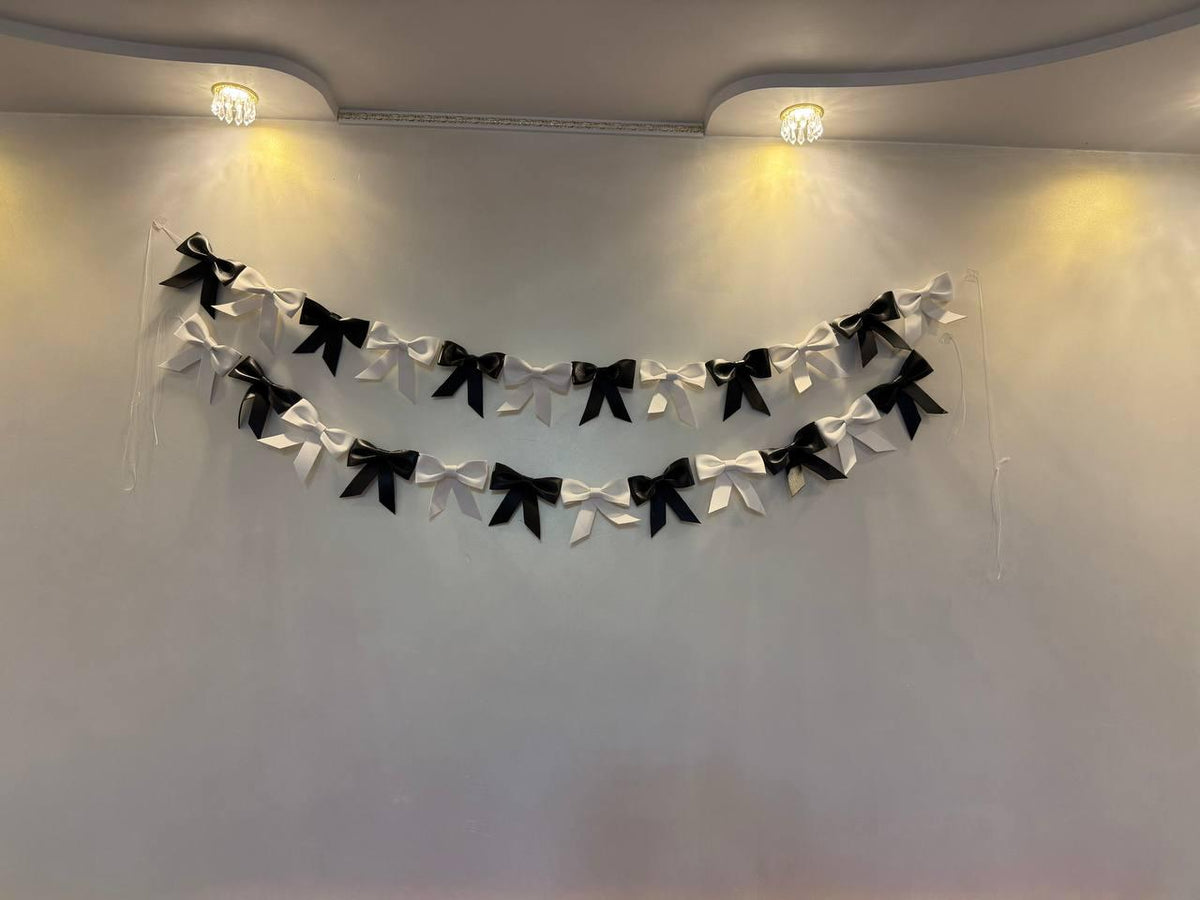 May include: Two strands of decorative bows in alternating black and white colors are suspended against a white wall. The bows are evenly spaced and create a festive, garland-like appearance. The lighting is soft, highlighting the bows' textures.