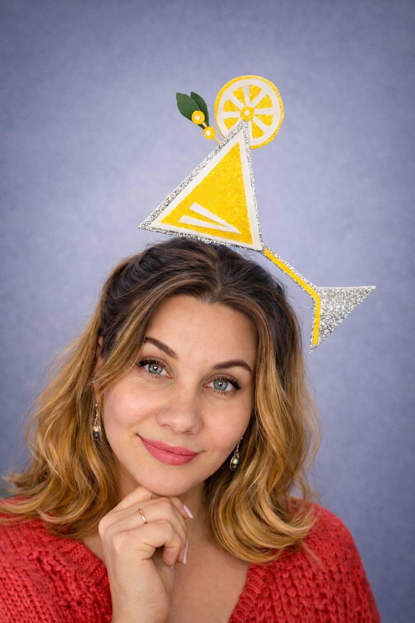 May include: A yellow and silver cocktail-themed headpiece with a lemon slice and green leaves. The headpiece is worn by a person with long, wavy blonde hair and a red sweater. The background is a solid light purple.