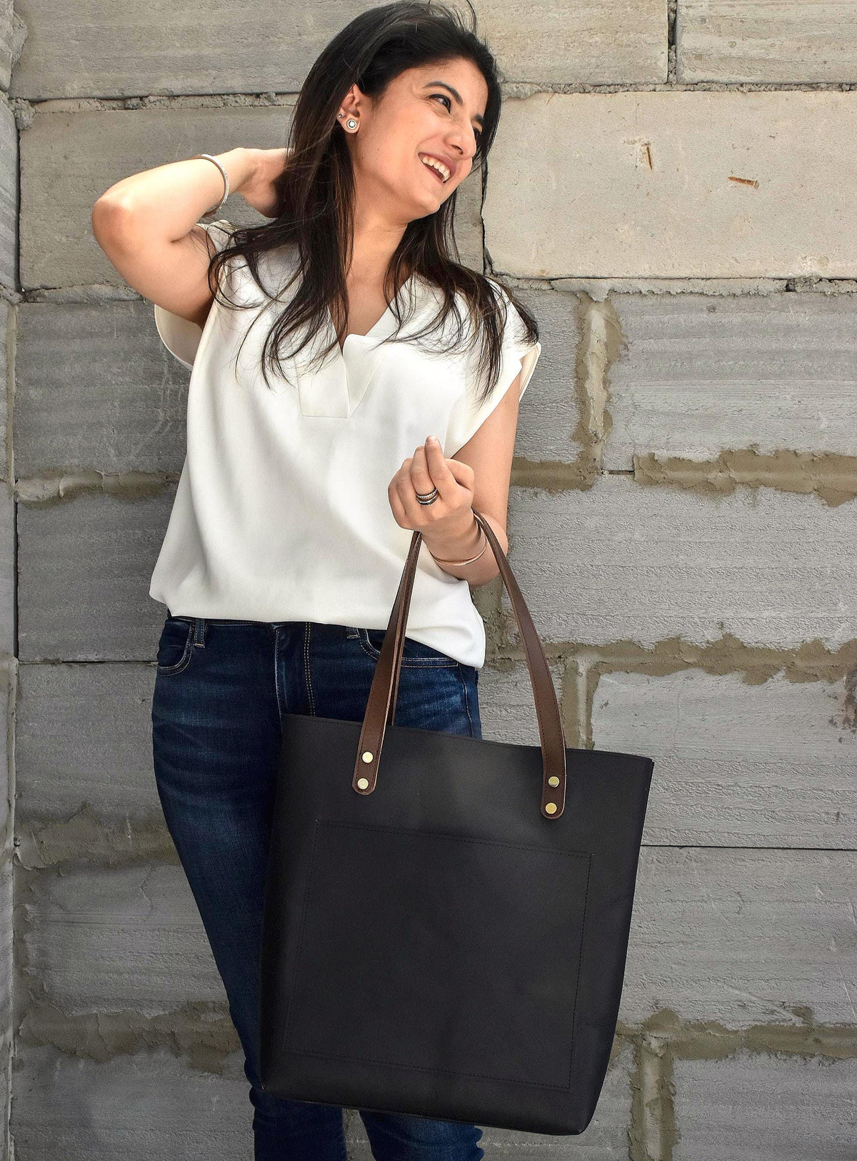 May include: Black tote bag with brown leather handles and gold rivets. The bag has a rectangular shape with a front pocket. The handles are attached with gold rivets. The bag is held by a person wearing a white top and blue jeans.