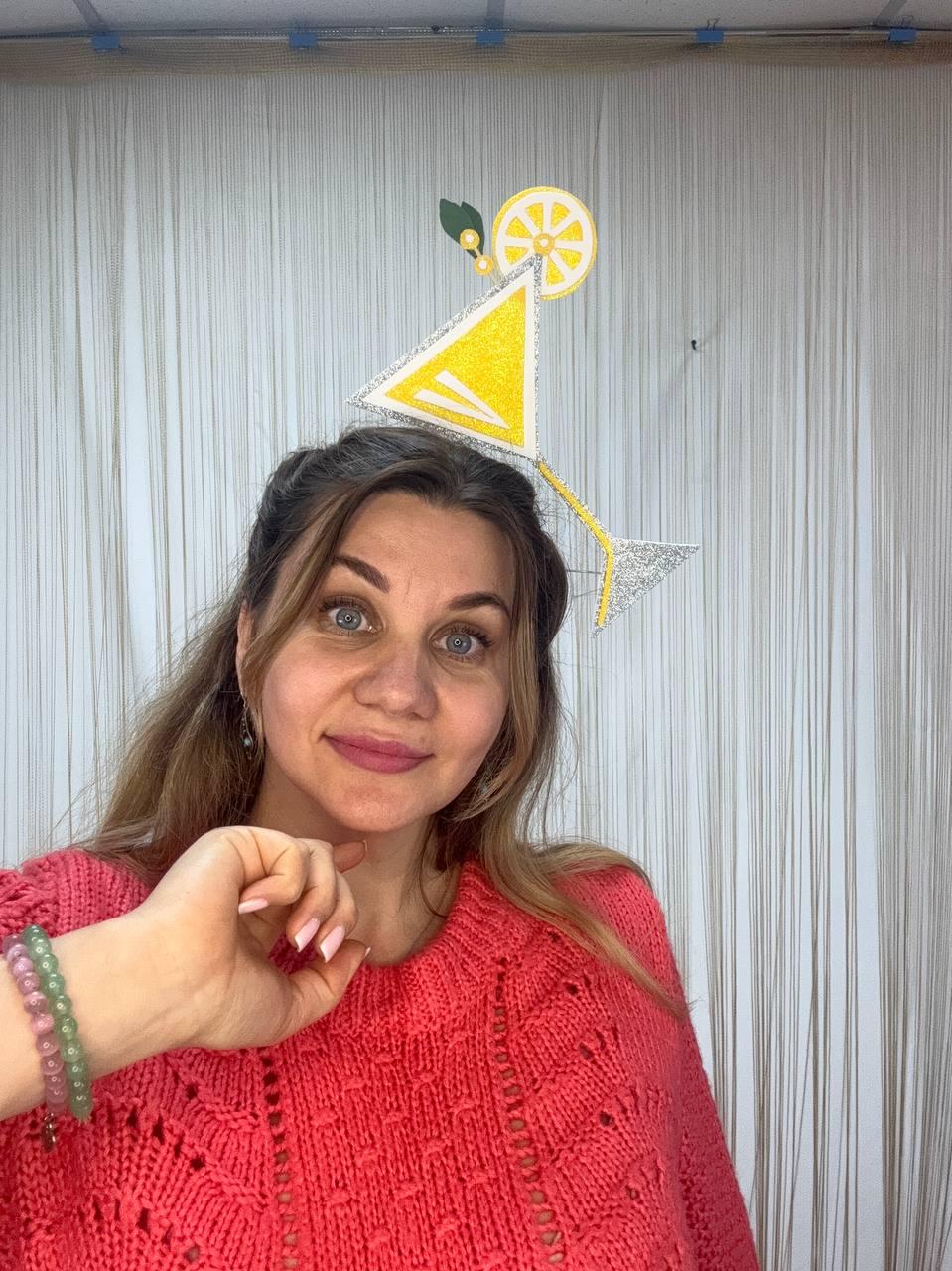 May include: A woman wearing a decorative headpiece shaped like a cocktail. The headpiece features a yellow triangle, a lemon slice, and a silver stem. She is wearing a coral-colored sweater and a bracelet.