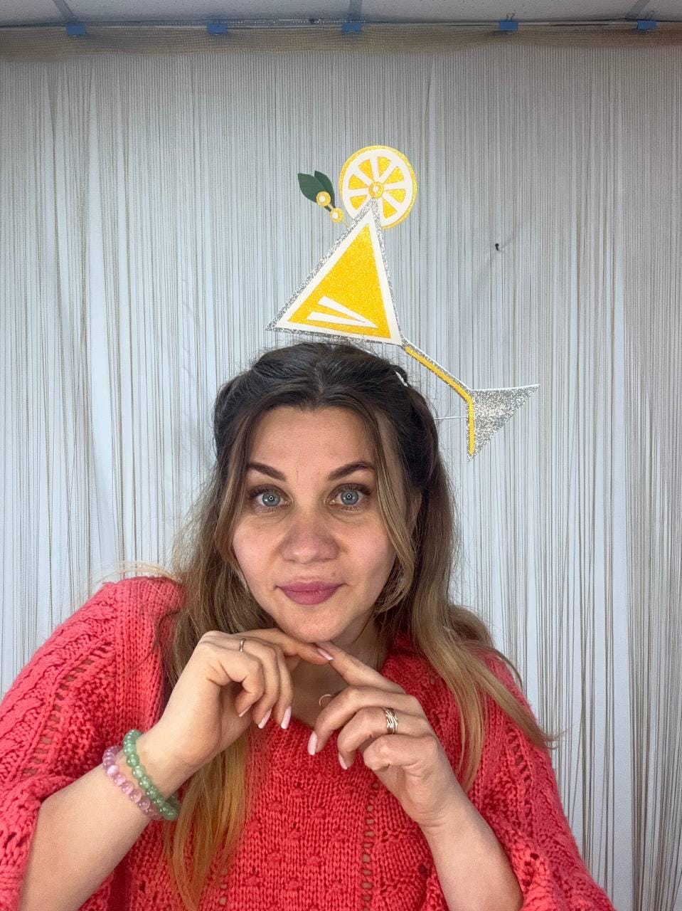 May include: A woman wearing a red sweater and a cocktail-themed headpiece. The headpiece features a yellow triangle, a lemon slice, and a silver stem. The background is a white curtain.