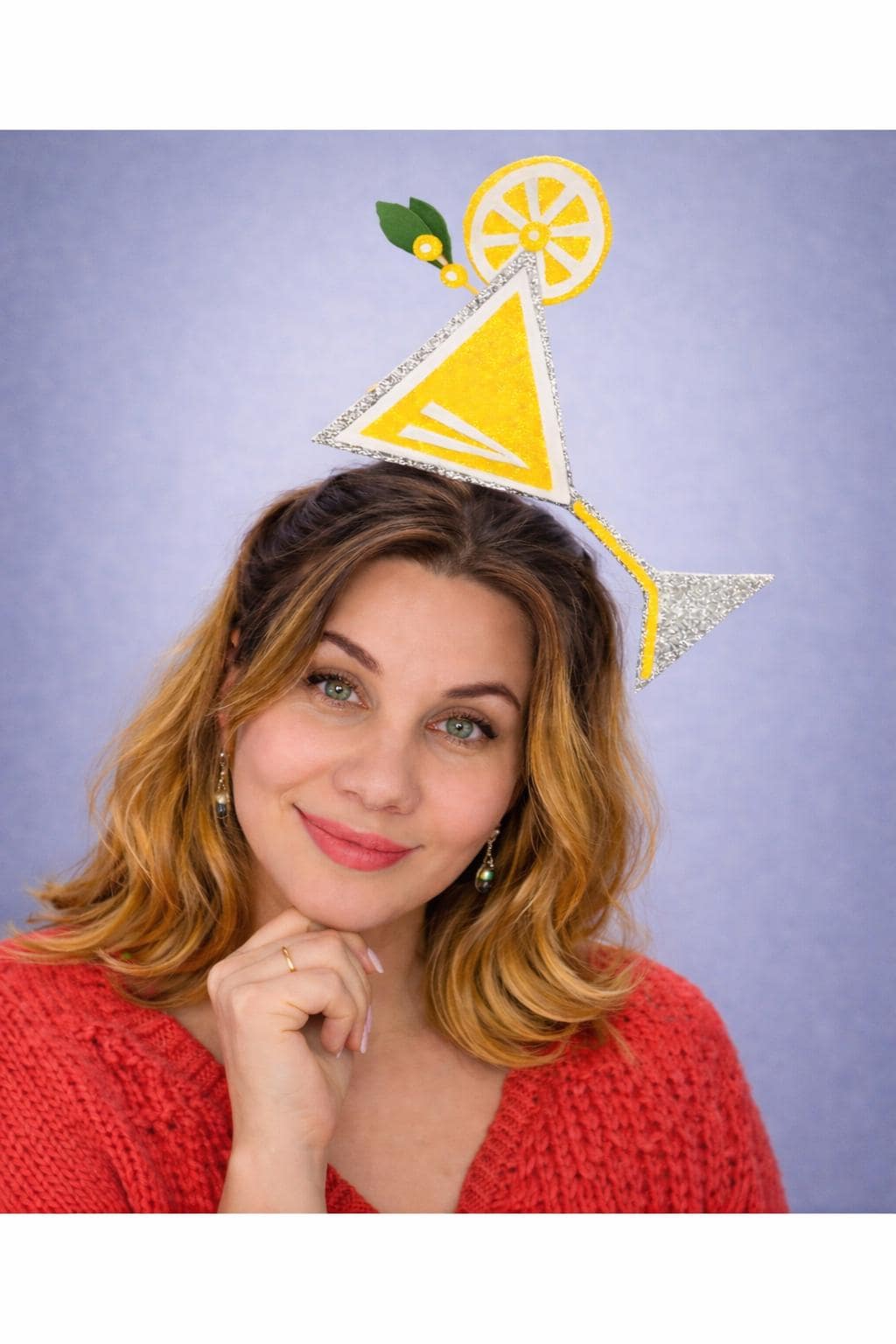 May include: A yellow and silver martini glass headpiece with a lemon slice and green leaves. The headpiece is worn by a person wearing a red sweater. The background is a solid light purple.