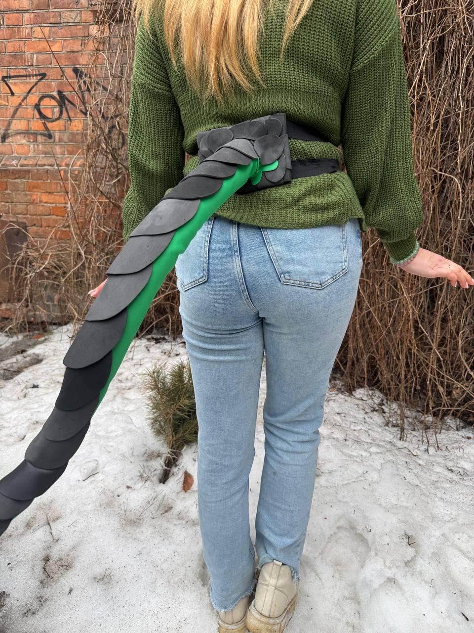 May include: A black and green dragon tail costume attached to a black belt. The tail is made of layered, dark gray scales over a green base. The costume is worn with a green sweater and blue jeans.