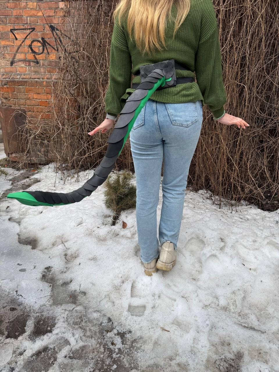 May include: A black and gray dragon tail costume with green accents, attached to a black belt. The tail is made of layered, scale-like material. The costume is worn with a green sweater and blue jeans.