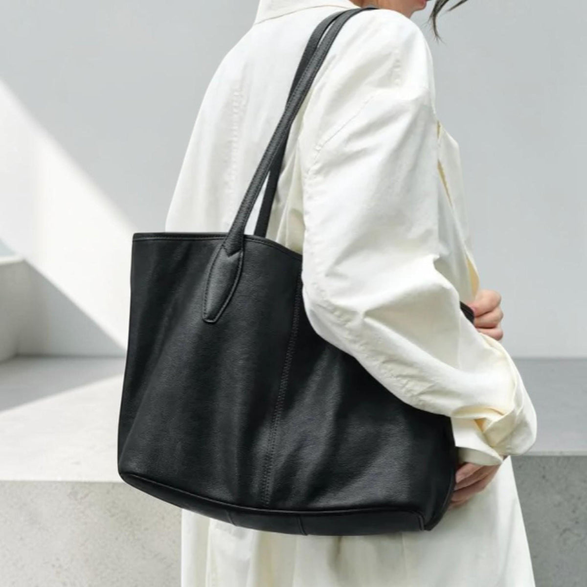 May include: A black leather tote bag with two long handles. The bag is rectangular with a structured design and a smooth texture. The bag is being carried by a person wearing a white shirt.
