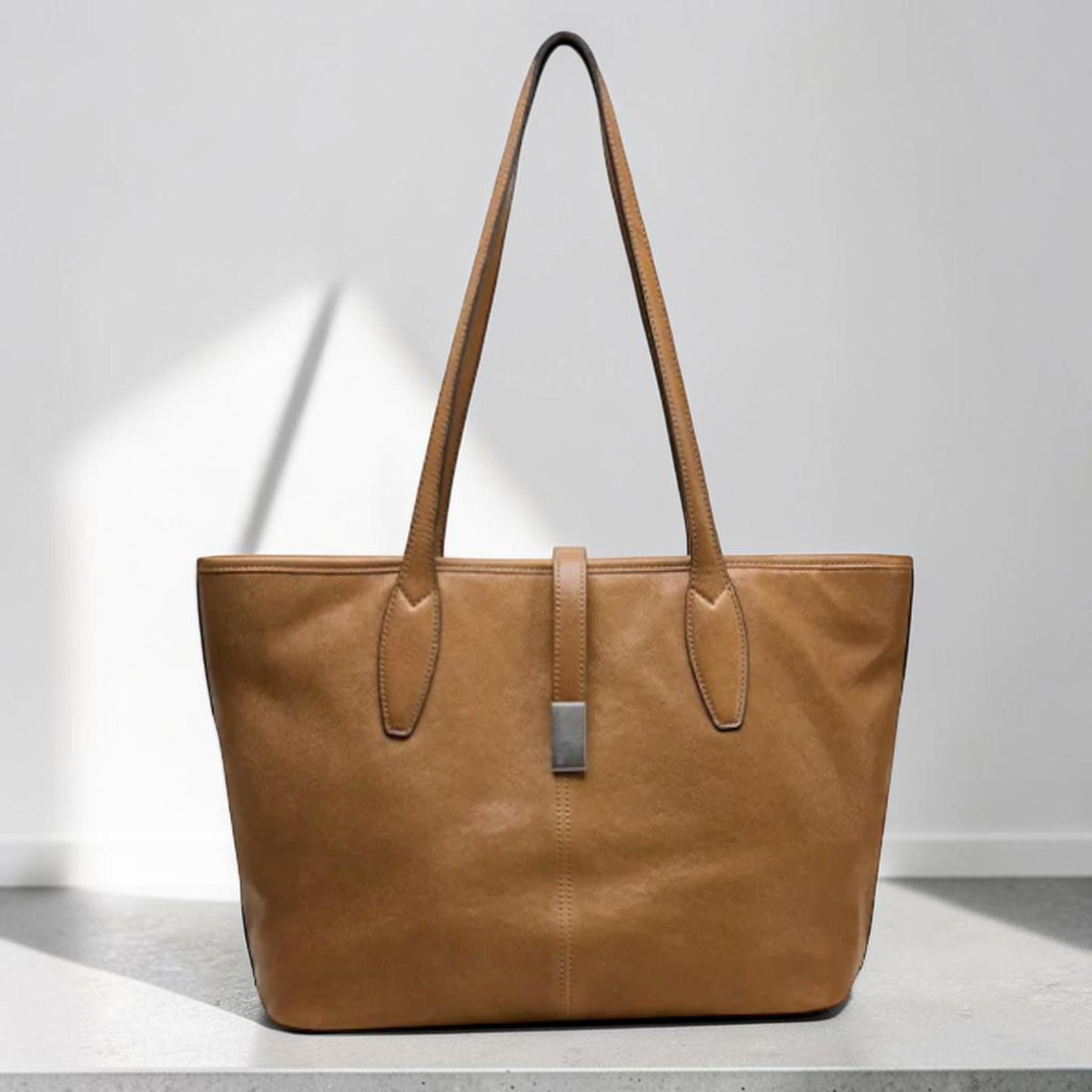 May include: A tan leather tote bag with long handles and a rectangular shape. The bag features a silver-colored clasp and a vertical strap closure. The bag is sitting on a white surface.