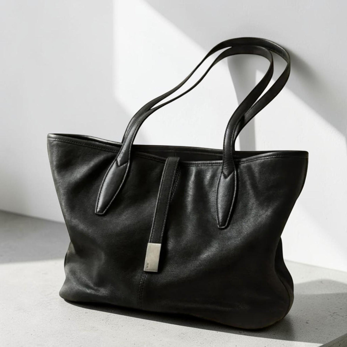 May include: A black leather tote bag with two long handles and a rectangular silver-colored clasp. The bag has a structured shape and appears to be made of a soft, supple leather. The bag is suitable for everyday use.