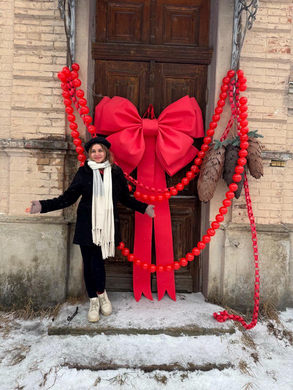 May include: A large red bow and matching garland decorate a wooden door. The garland is made of red spheres and pinecones. The door is framed by a light-colored brick wall. A person is standing in front of the door.