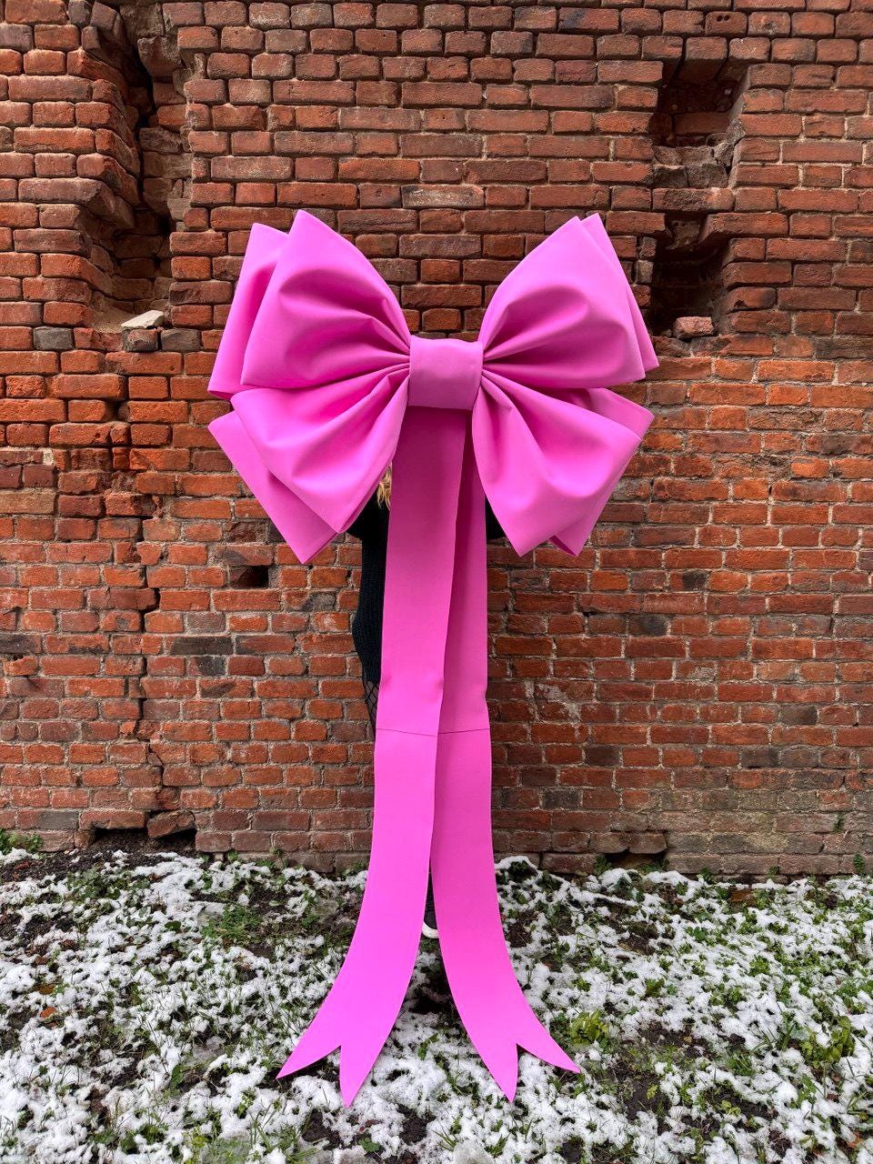 May include: A large, bright pink bow with long, flowing ribbons. The bow is made of a smooth material and is set against a weathered brick wall. The ribbons extend downwards, adding to the bow's dramatic appearance.