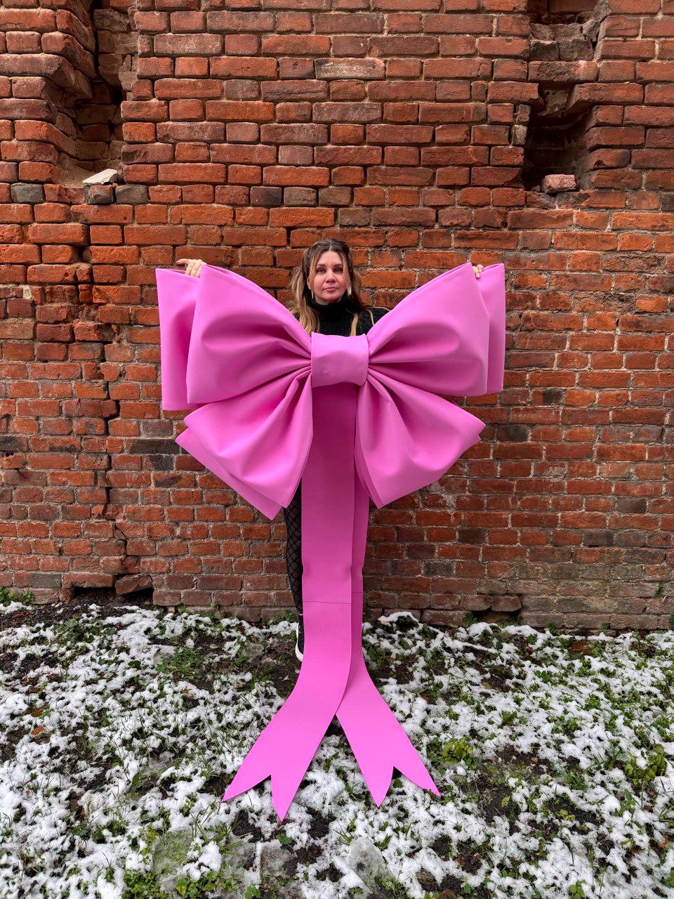 May include: A large, vibrant pink bow with long, flowing tails. The bow is held in front of a brick wall, creating a striking contrast. The bow's size and color make it a bold decorative element.