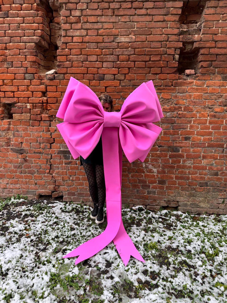 May include: A large, bright pink bow with long, flowing ribbons. The bow is made of a smooth material and is positioned in front of a brick wall. The bow's size suggests it could be used for decoration or as a prop.