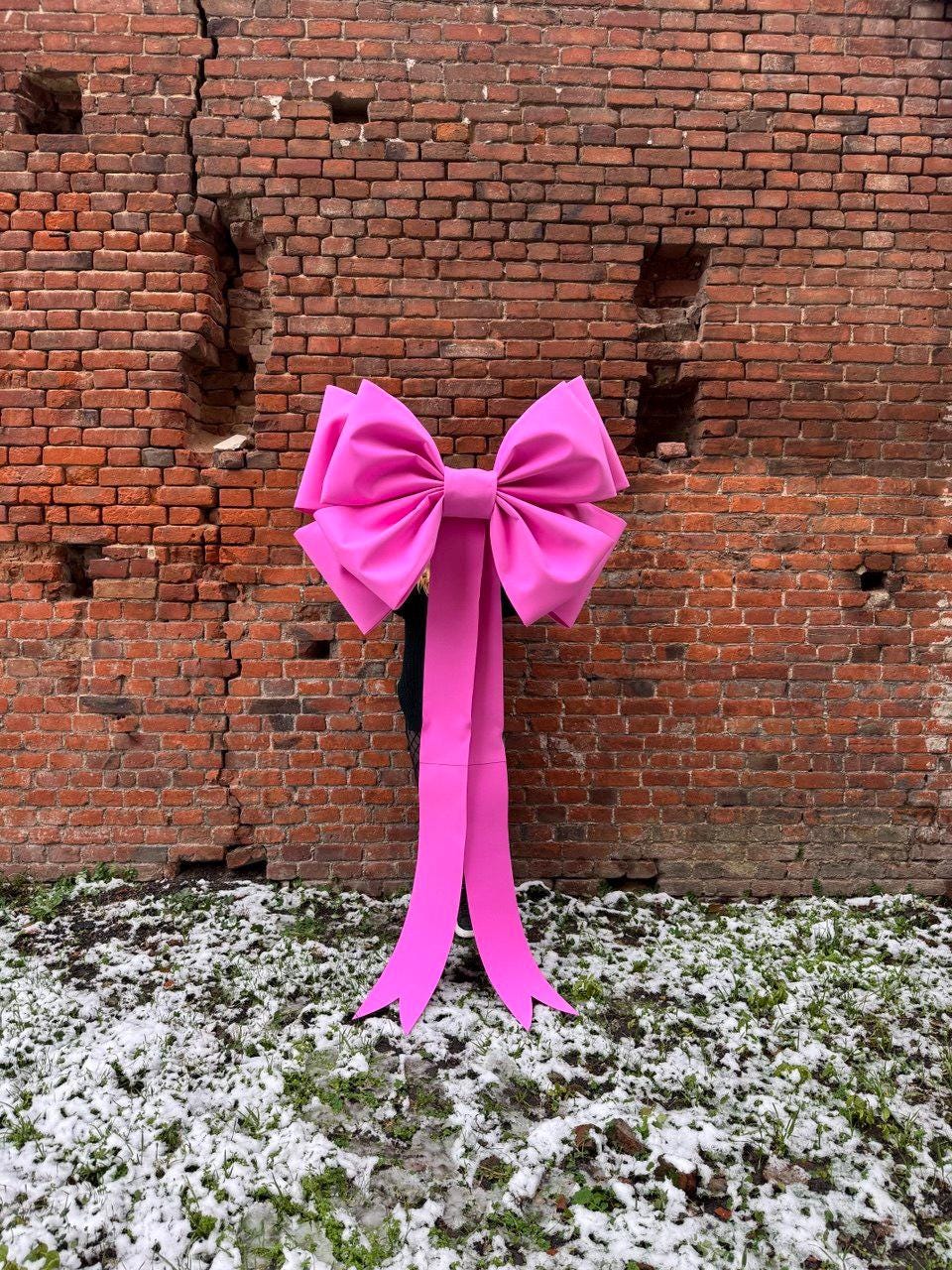 May include: A large, bright pink bow with long, flowing streamers. The bow is made of a smooth, possibly satin-like material. It is set against a weathered brick wall and a snowy ground.