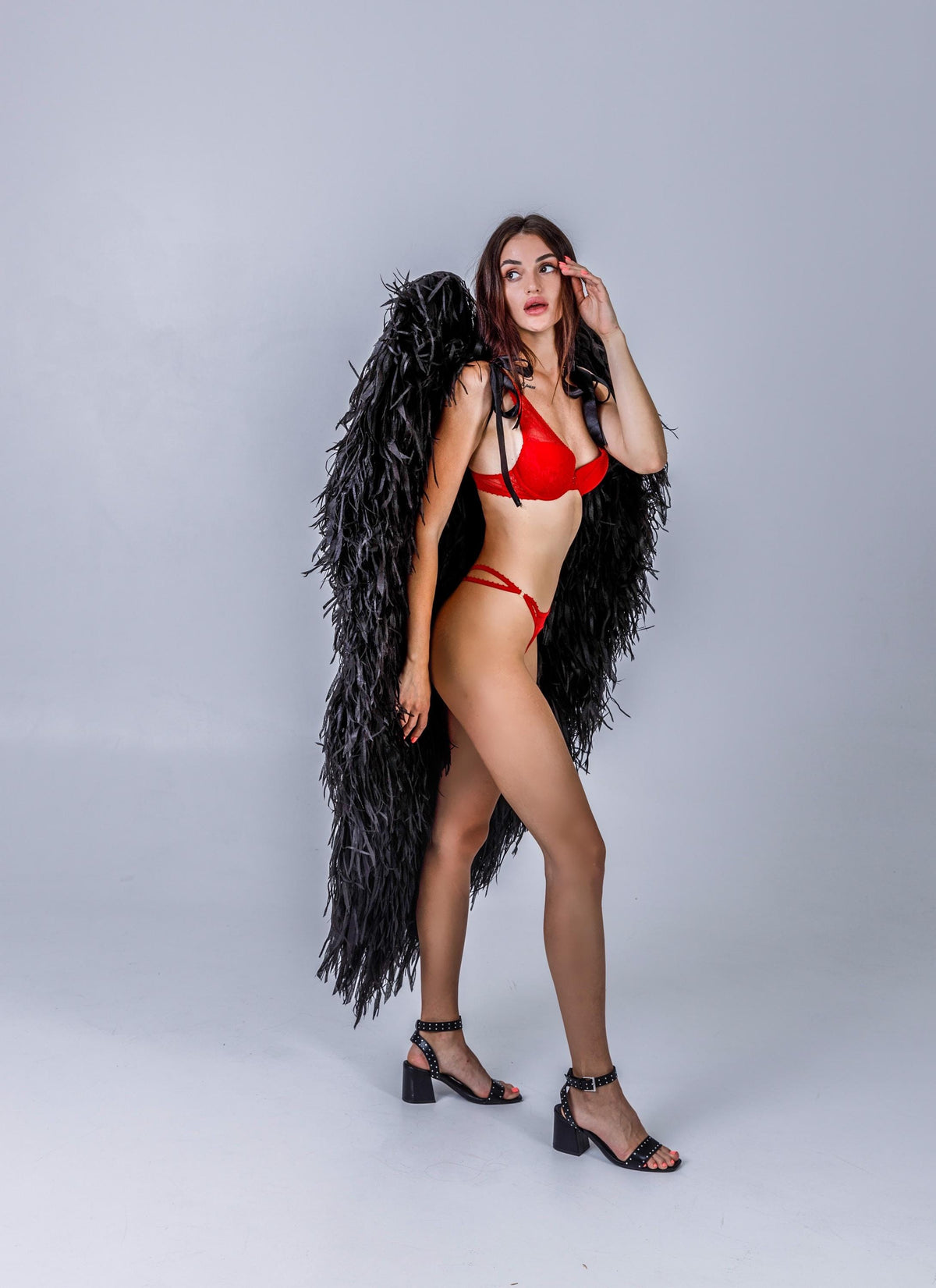 May include: A person wearing a red lingerie set and black feathered angel wings. The person is also wearing black heeled sandals with studded details. The background is a solid light gray.