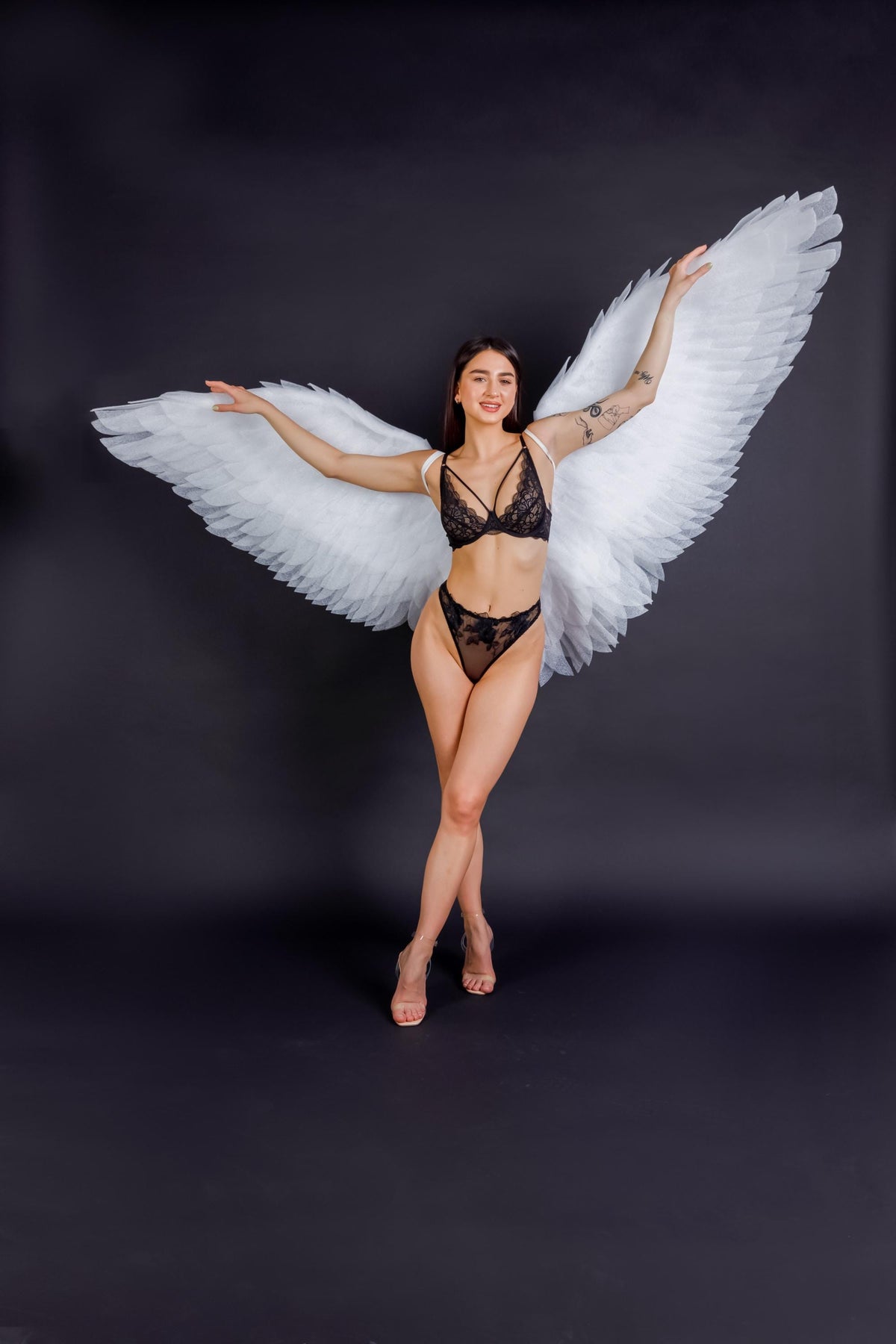 May include: A woman wearing black lingerie and large white angel wings poses against a dark background. The lingerie features delicate lace details. The wings are large and feathered, creating a dramatic effect. The woman is wearing clear heels.