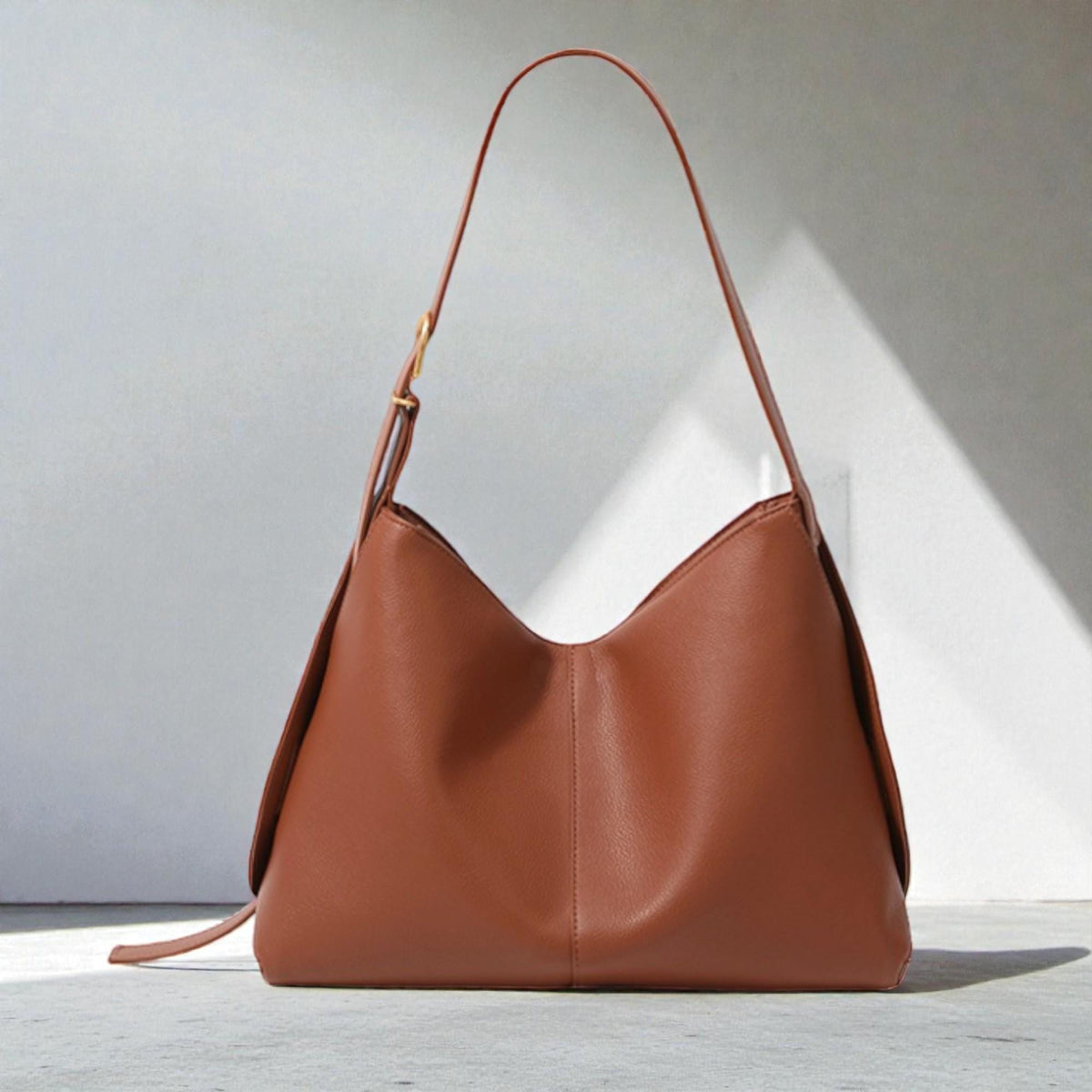May include: A brown leather shoulder bag with a single strap and a gold-colored buckle. The bag has a curved shape and a minimalist design, suitable for everyday use. The bag is sitting on a white surface.