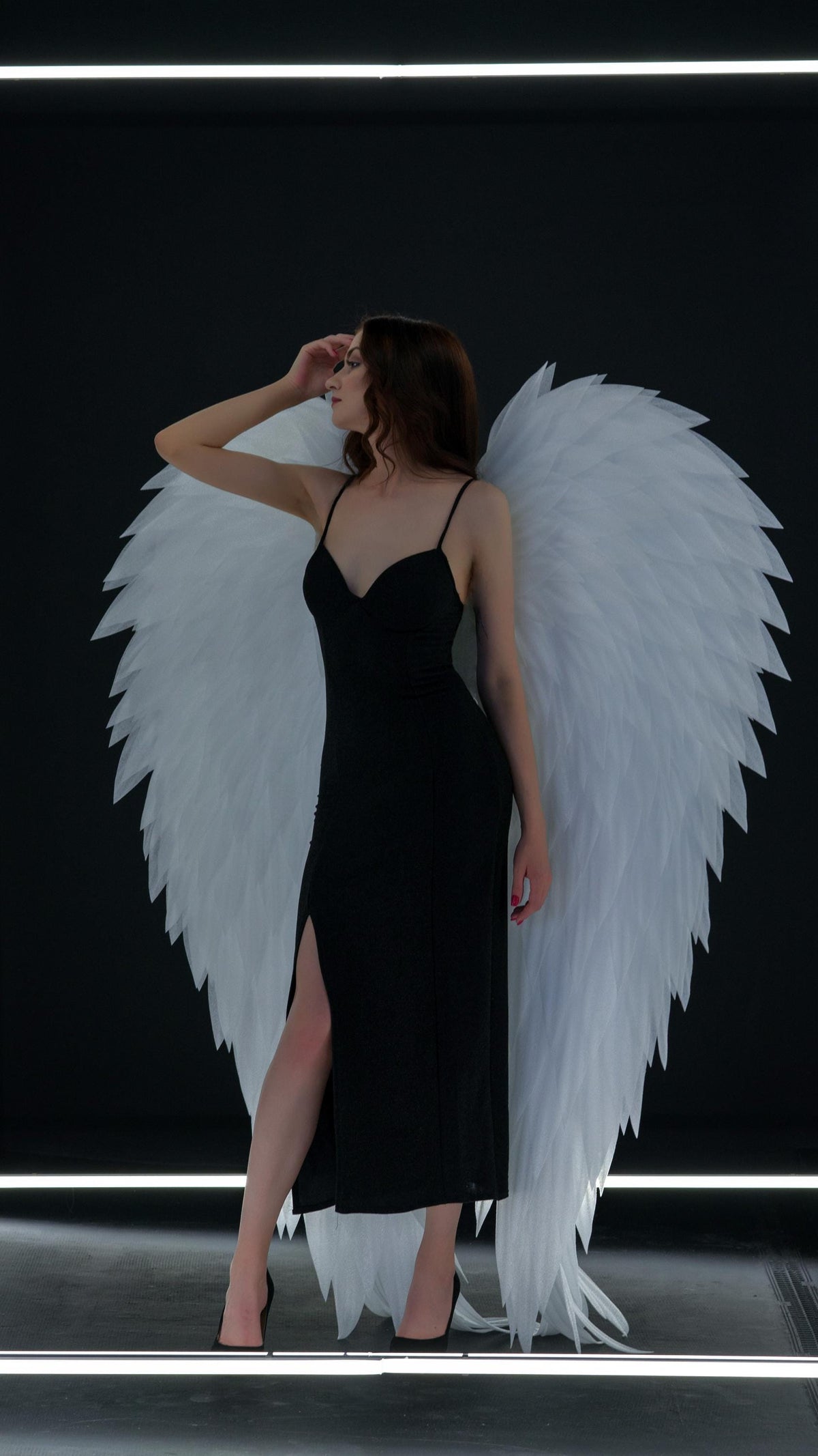 May include: A woman wearing a black dress with a high slit and black heels, posing with large white feathered angel wings against a black background. The dress has thin straps.