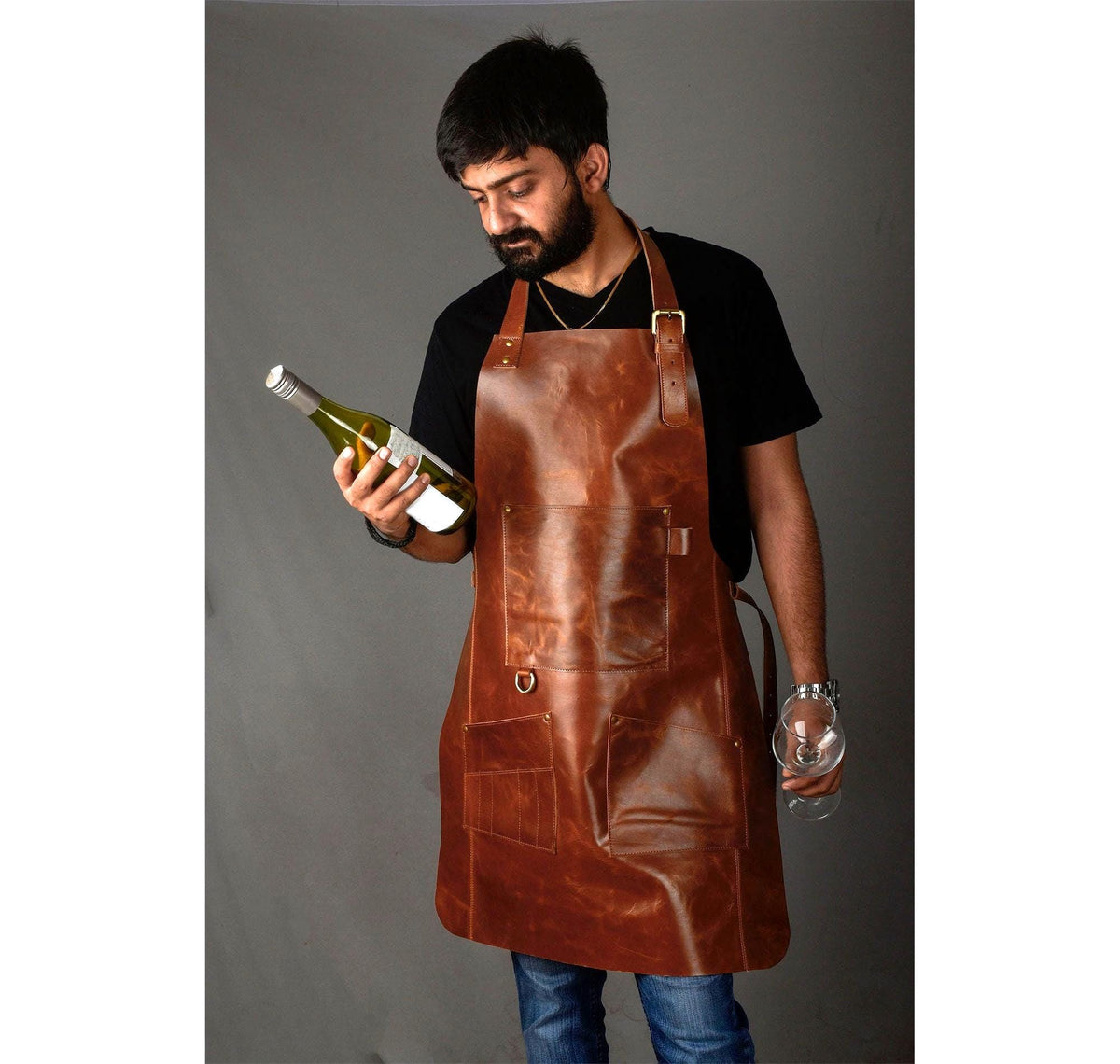 May include: A brown leather apron with multiple pockets and adjustable straps. The apron is worn by a person holding a wine bottle and a wine glass. The apron has a classic design and is made of a durable material.