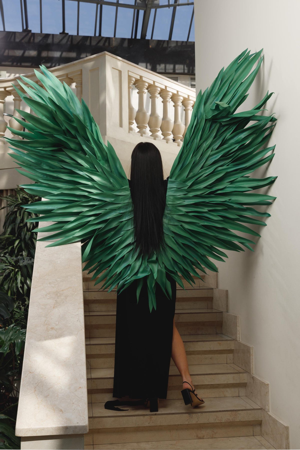 May include: A person wearing large, green, feathered wings and a black dress stands on a staircase. The wings are the focal point, with each feather-like element meticulously arranged. The background includes a white balustrade and a glass ceiling.