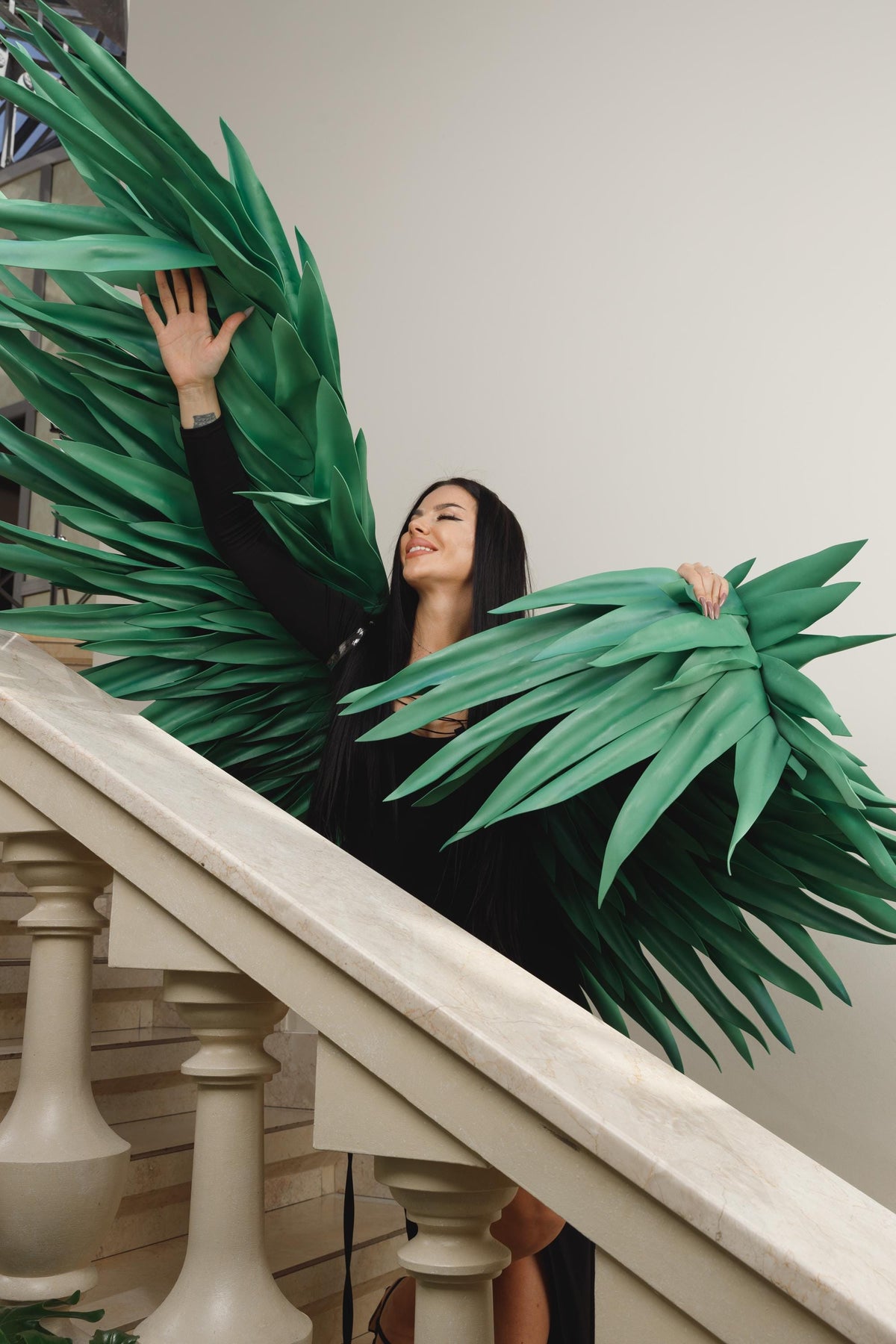 May include: A woman wearing a black dress and large, green, stylized wings, posing on a staircase. The wings are made of layered, pointed, leaf-like shapes. The staircase has a white balustrade.