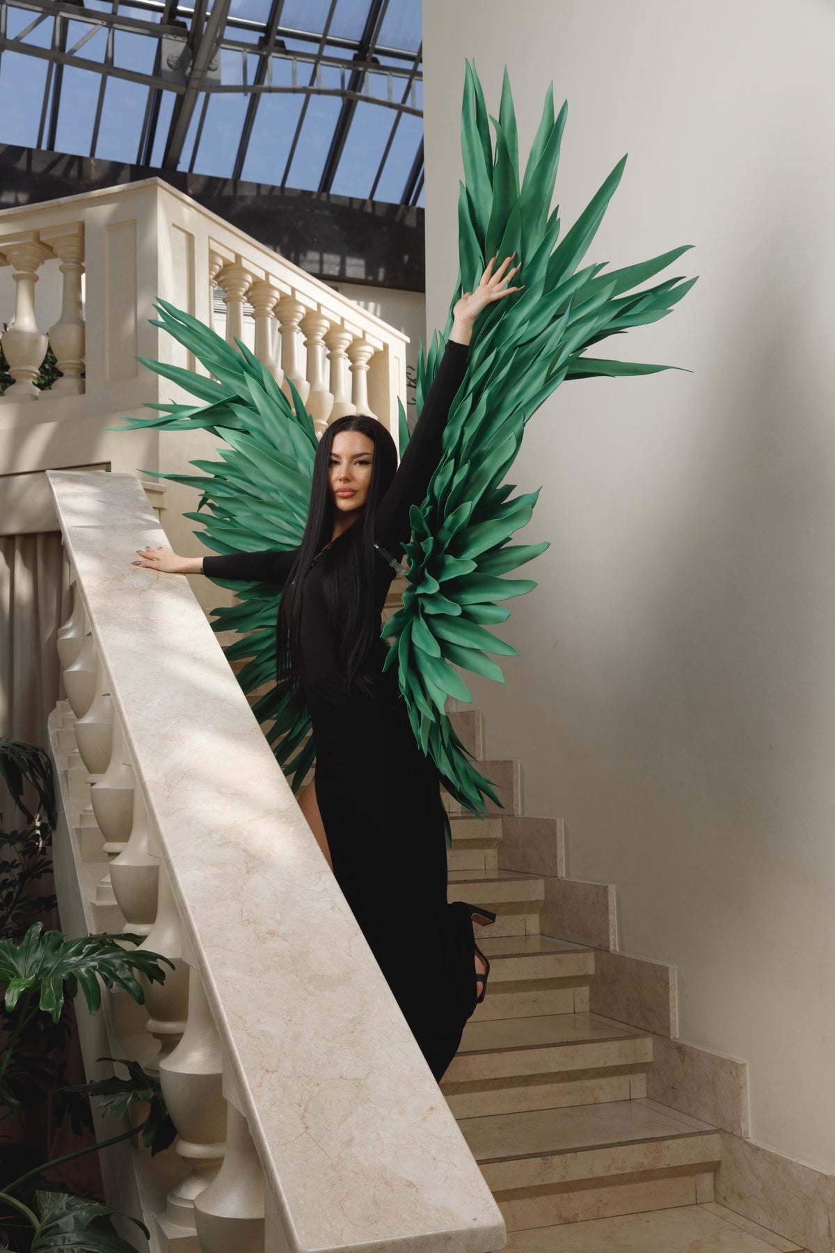 May include: A woman in a black dress and long sleeves poses on a staircase, wearing large, green, stylized wings. The background includes a white balustrade, stairs, and a glass ceiling, creating a dramatic and artistic composition.