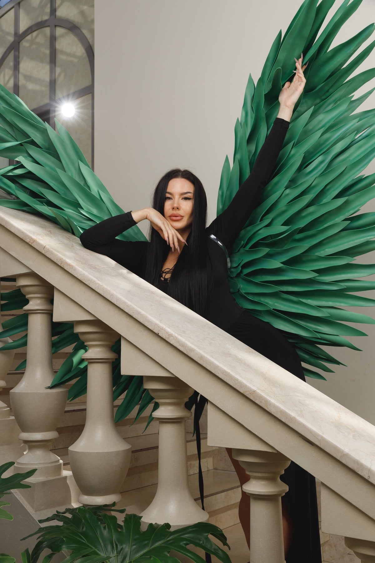 May include: A woman in a black dress poses on a staircase with large green paper wings. The wings are made of layered, pointed leaves. The staircase has white balusters and a marble handrail.