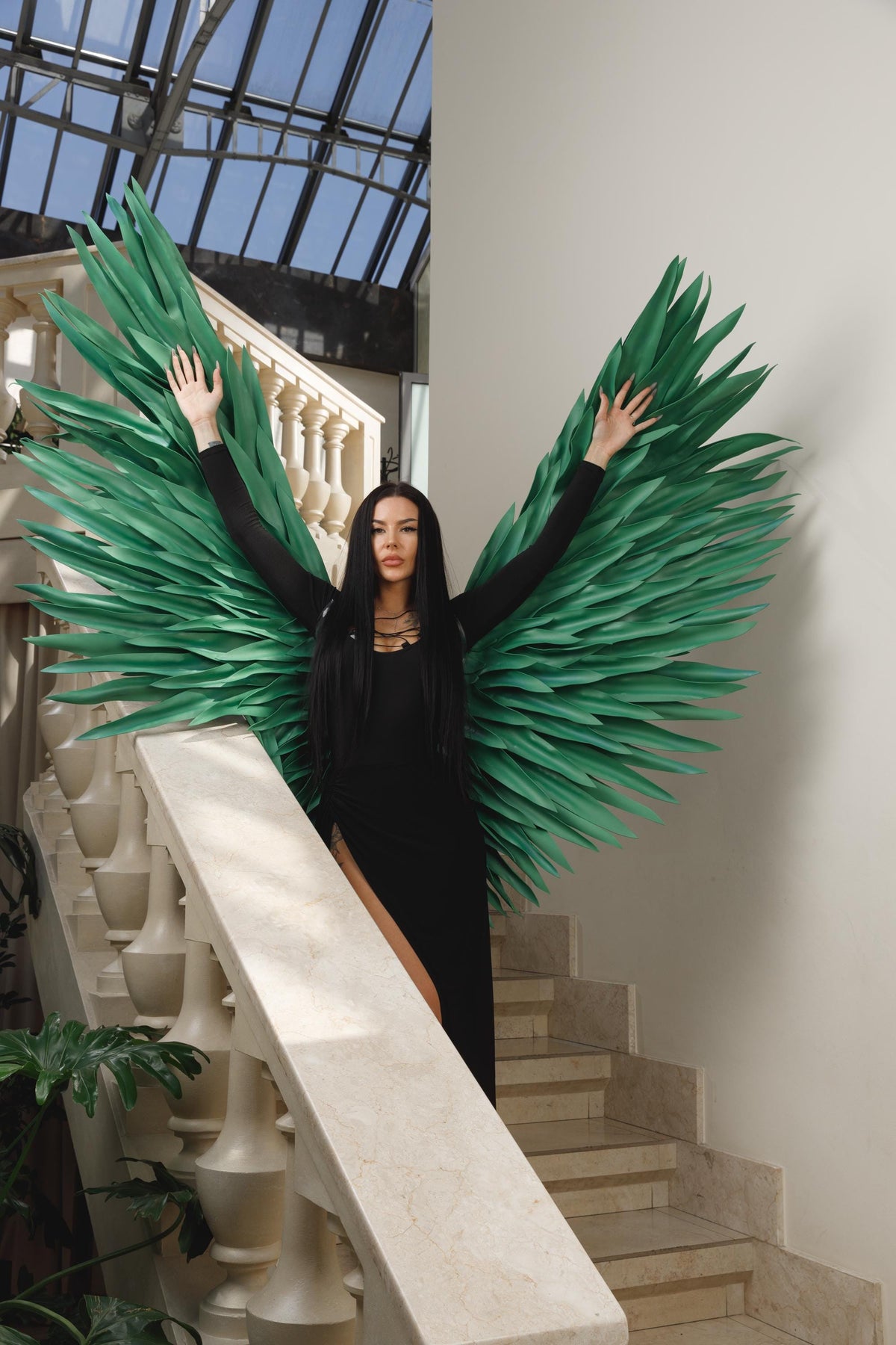 May include: A woman in a black dress with large, green, feathered wings, standing on a marble staircase. The wings are the focal point, with a layered design and a vibrant green color. The background includes a glass ceiling and white walls.